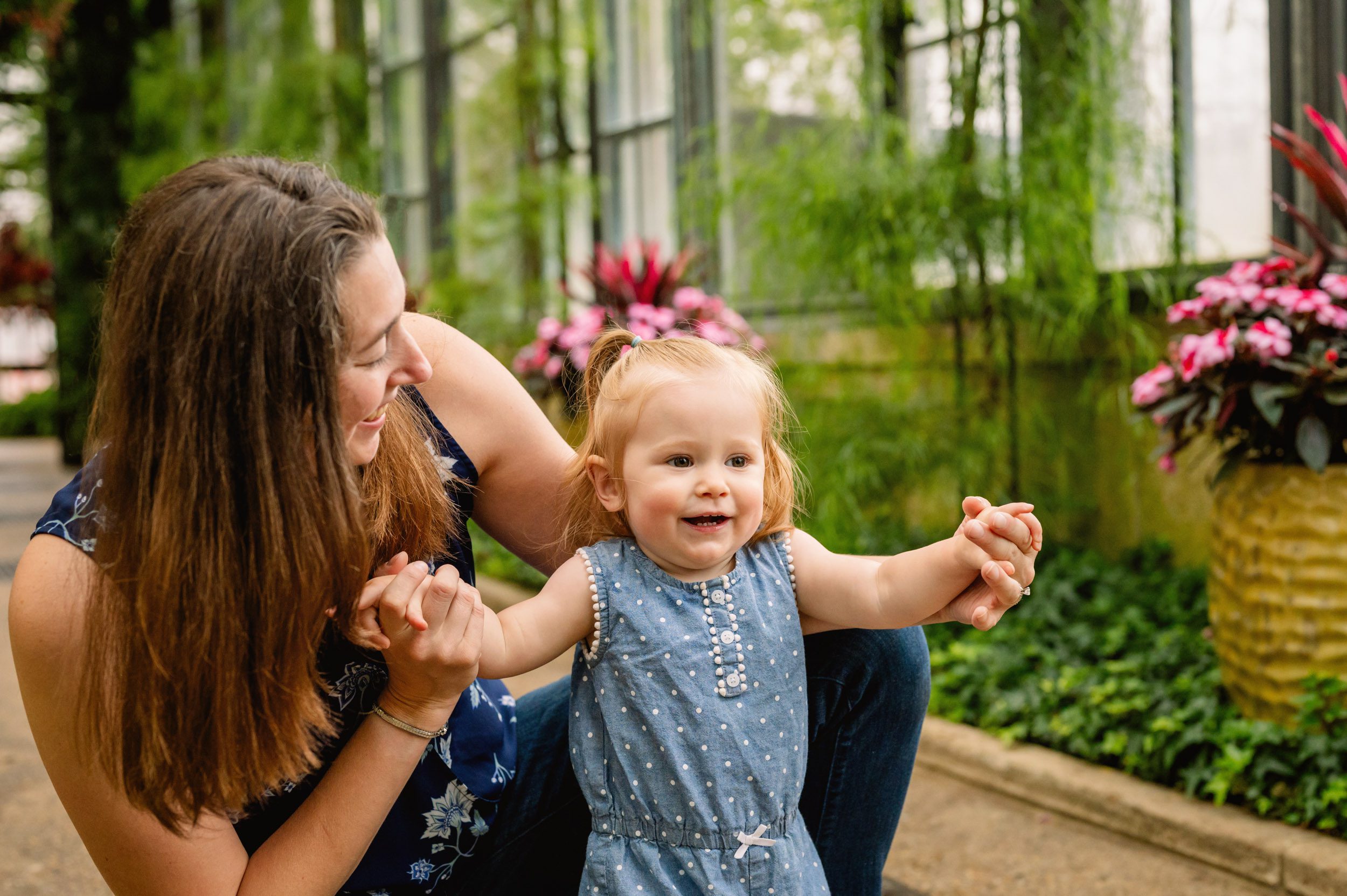 a mom kneeling down in a greenhouse and both holding her daughter's hands as she smiles at her during a fmaily photoshoot