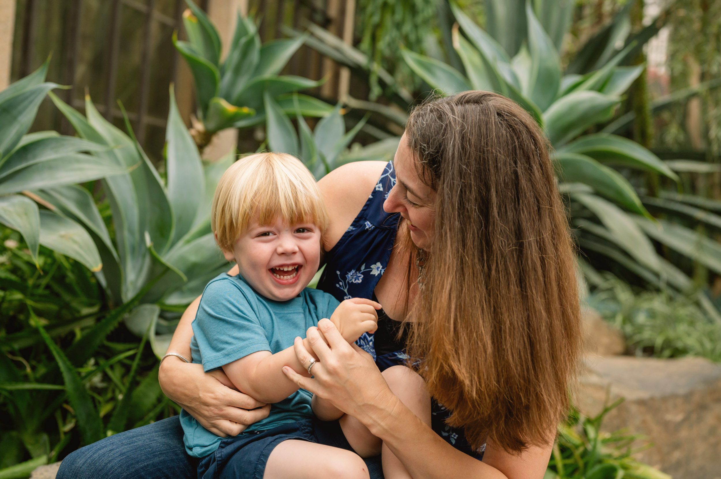 a mom sitting on a rock in a greenhouse with her young son on her lap and tickling him as he smiles at the camera during a family photo session