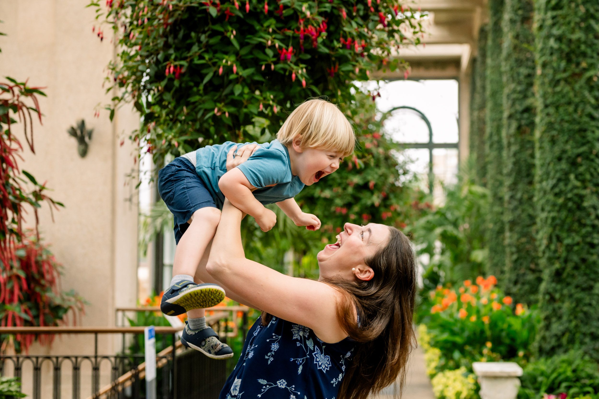 a mom standing in a greenhouse surrounded by colorful flowers in bloom as she lifts her son up in the air and they both laugh together during a family photography session