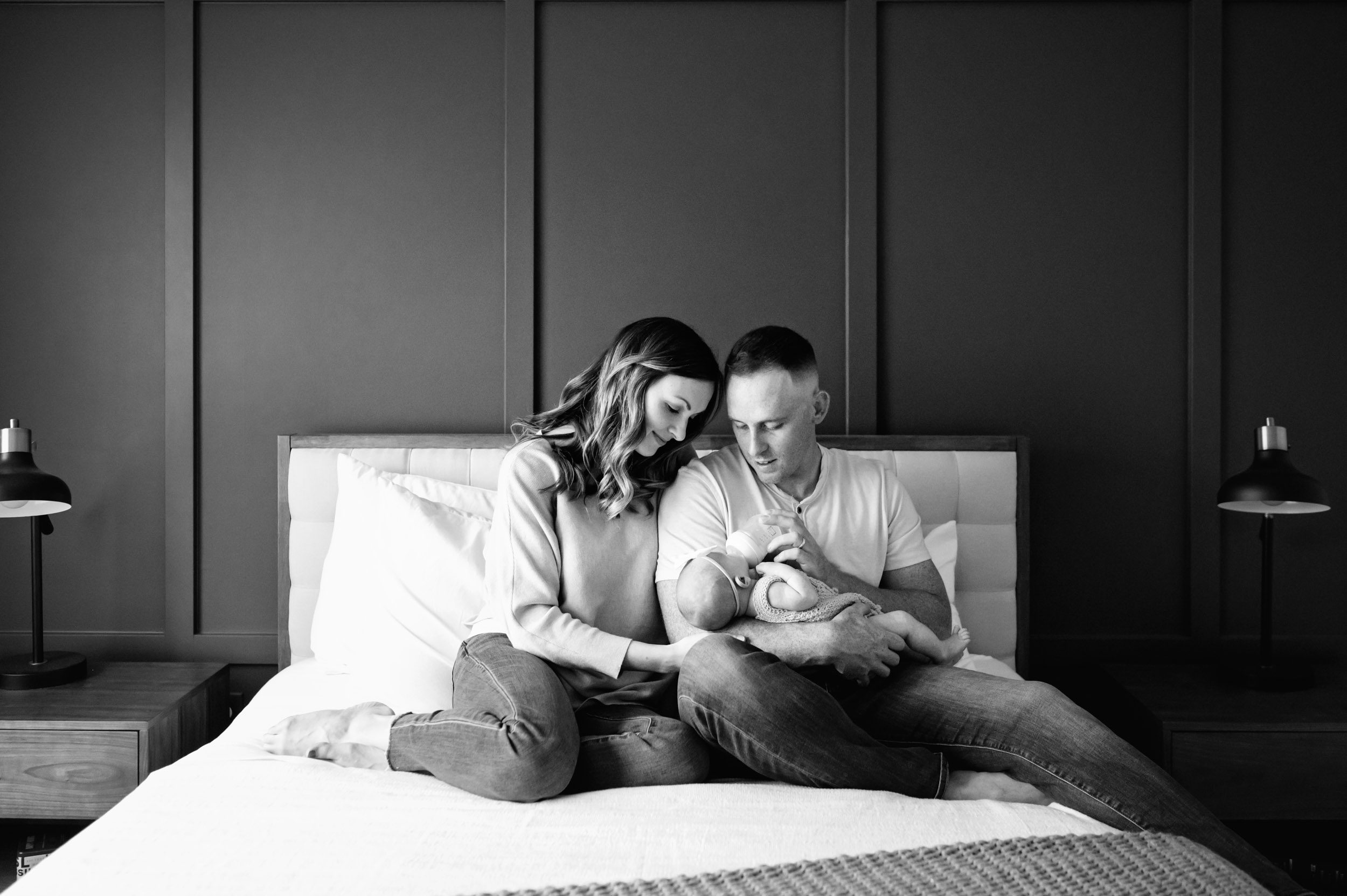 a black and white picture of new parents sitting on a bed holding their baby girl in their arms and smiling down at her while dad feeds the baby a bottle during an in home newborn photography session