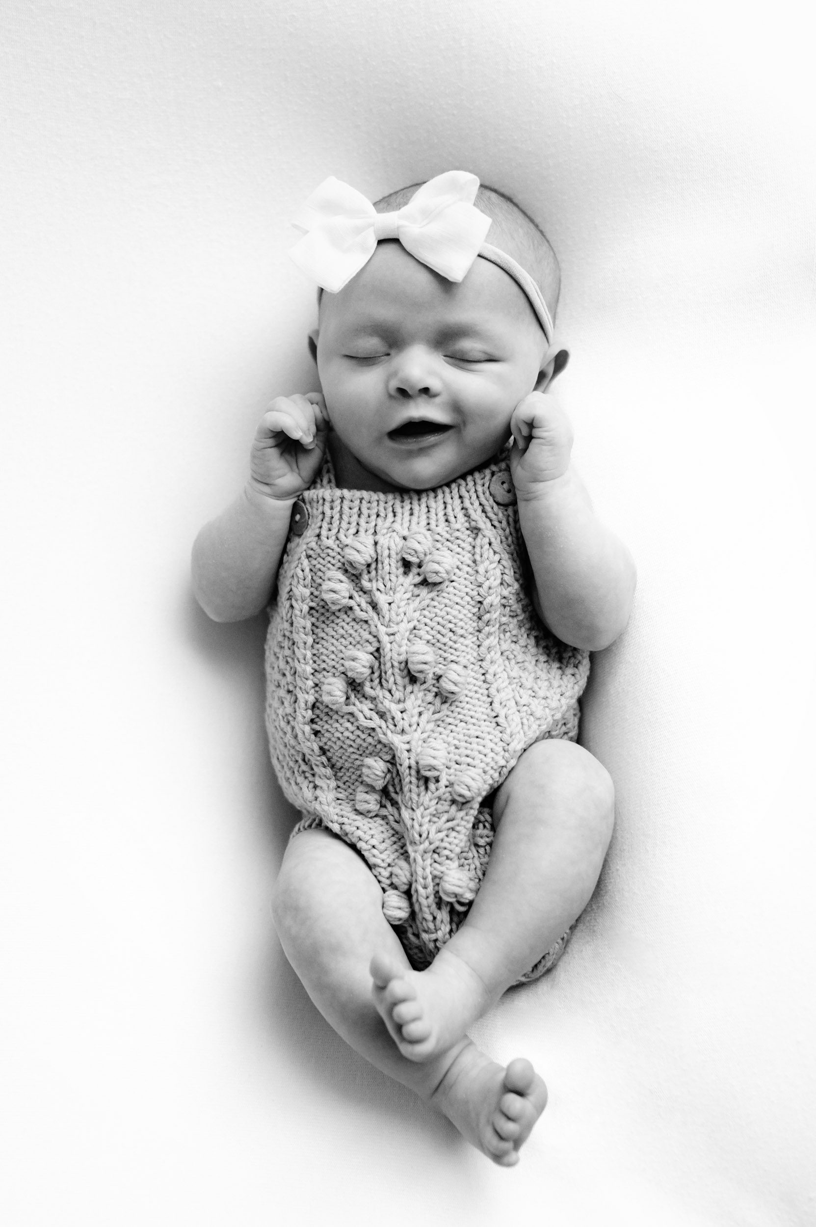 a black and white picture of a baby girl wearing a knit romper laying on a white background and smiling as she sleeps during a newborn photo session
