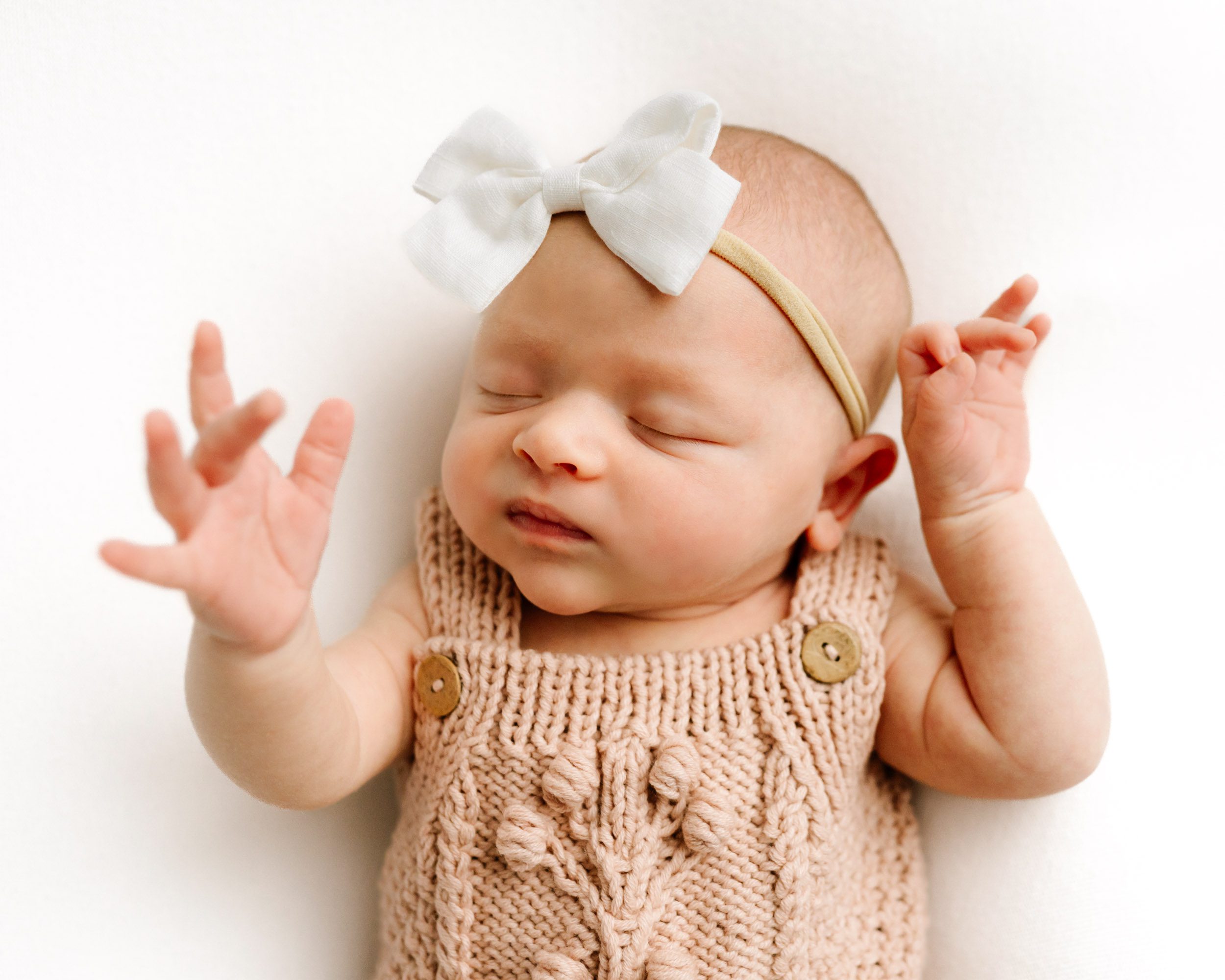 a baby girl wearing a pink knit romper and white headband laying on a white backdrop reaching her hand out toward the camera during a newborn photoshoot