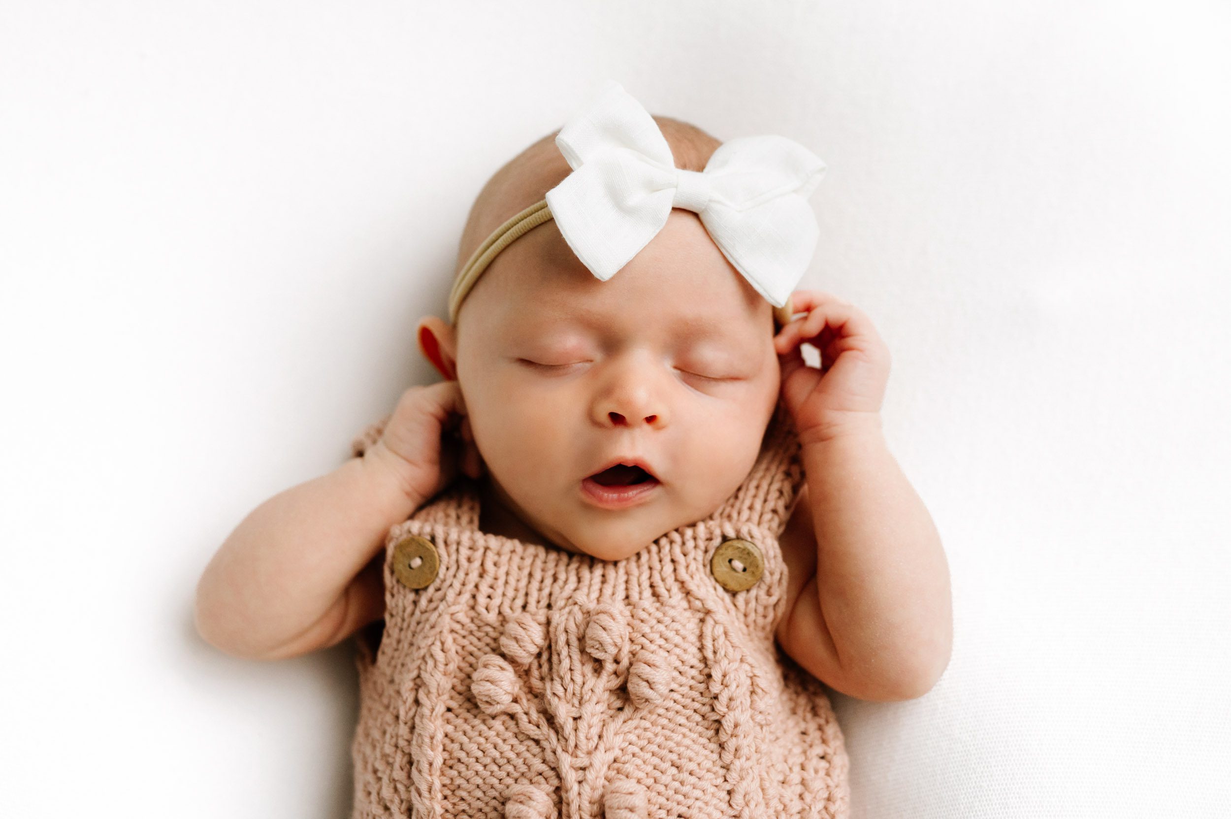 a baby girl wearing a pink knit romper and white headband laying on a white backdrop touching her hands to her cheeks with a surprised expression on her face during a newborn photoshoot