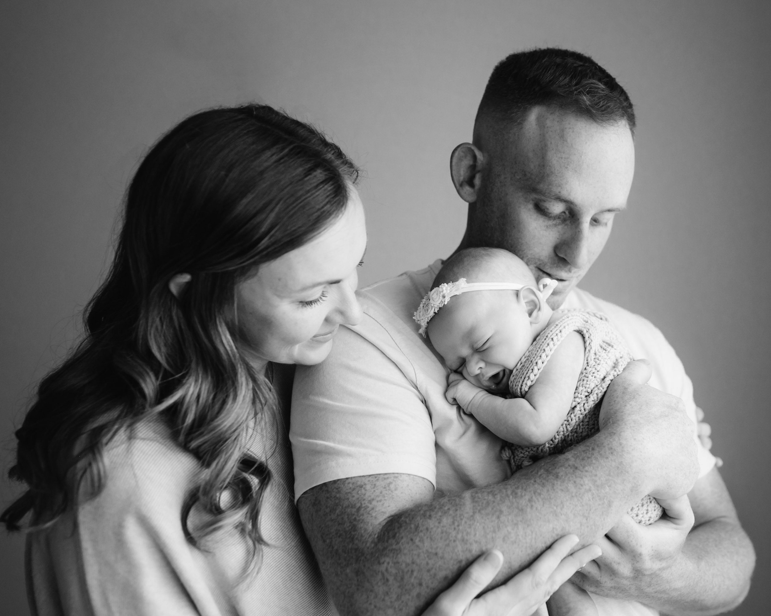 a black and white picture of a baby girl snuggled up against dad's chest sleeping with a big smile on her face while mom hugs dad from behind during a newborn photo session