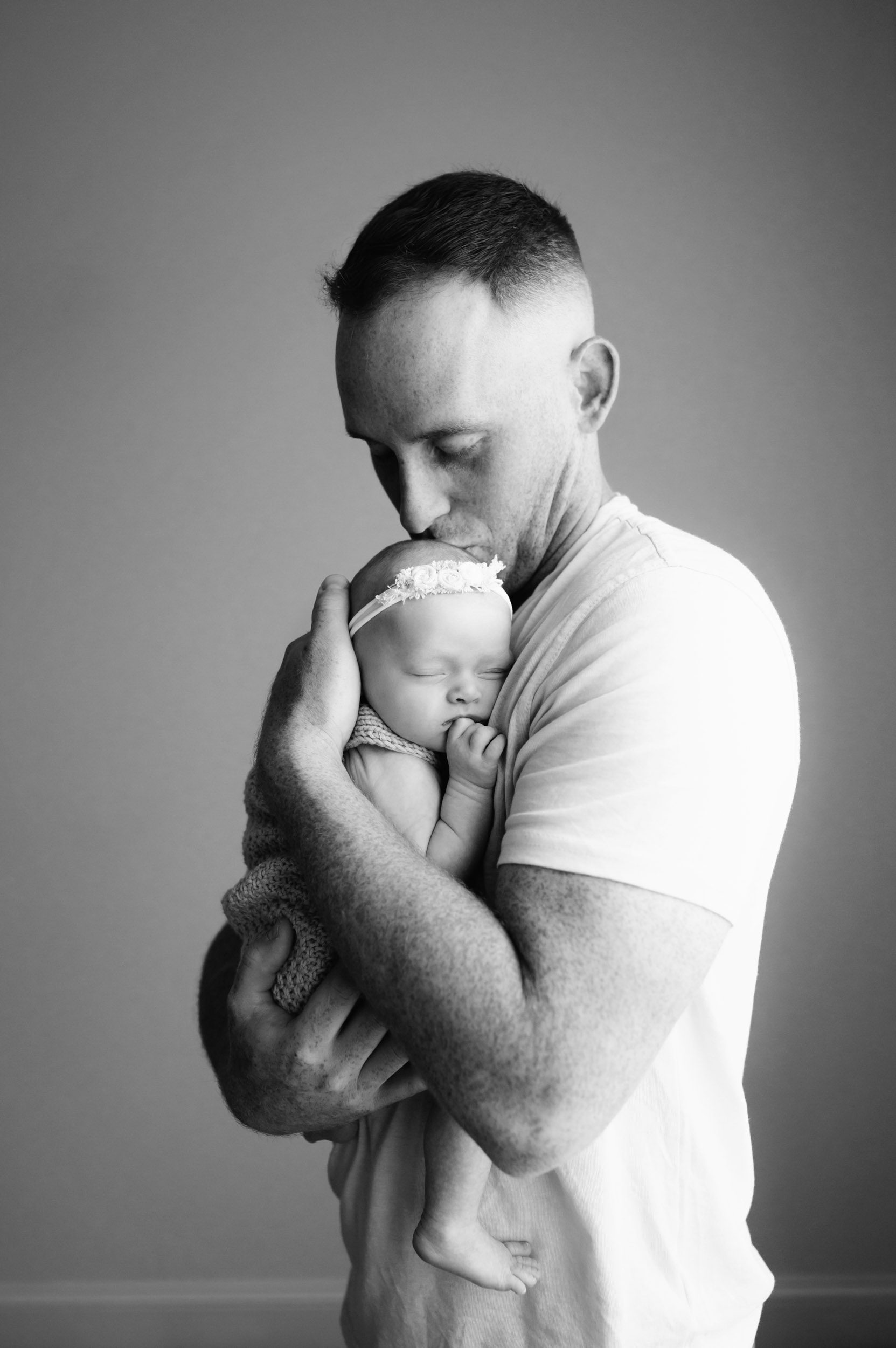 a black and white picture of a new father holding his baby girl against his chest as she sleeps peacefully and giving her a kiss on the head during an in home newborn photoshoot