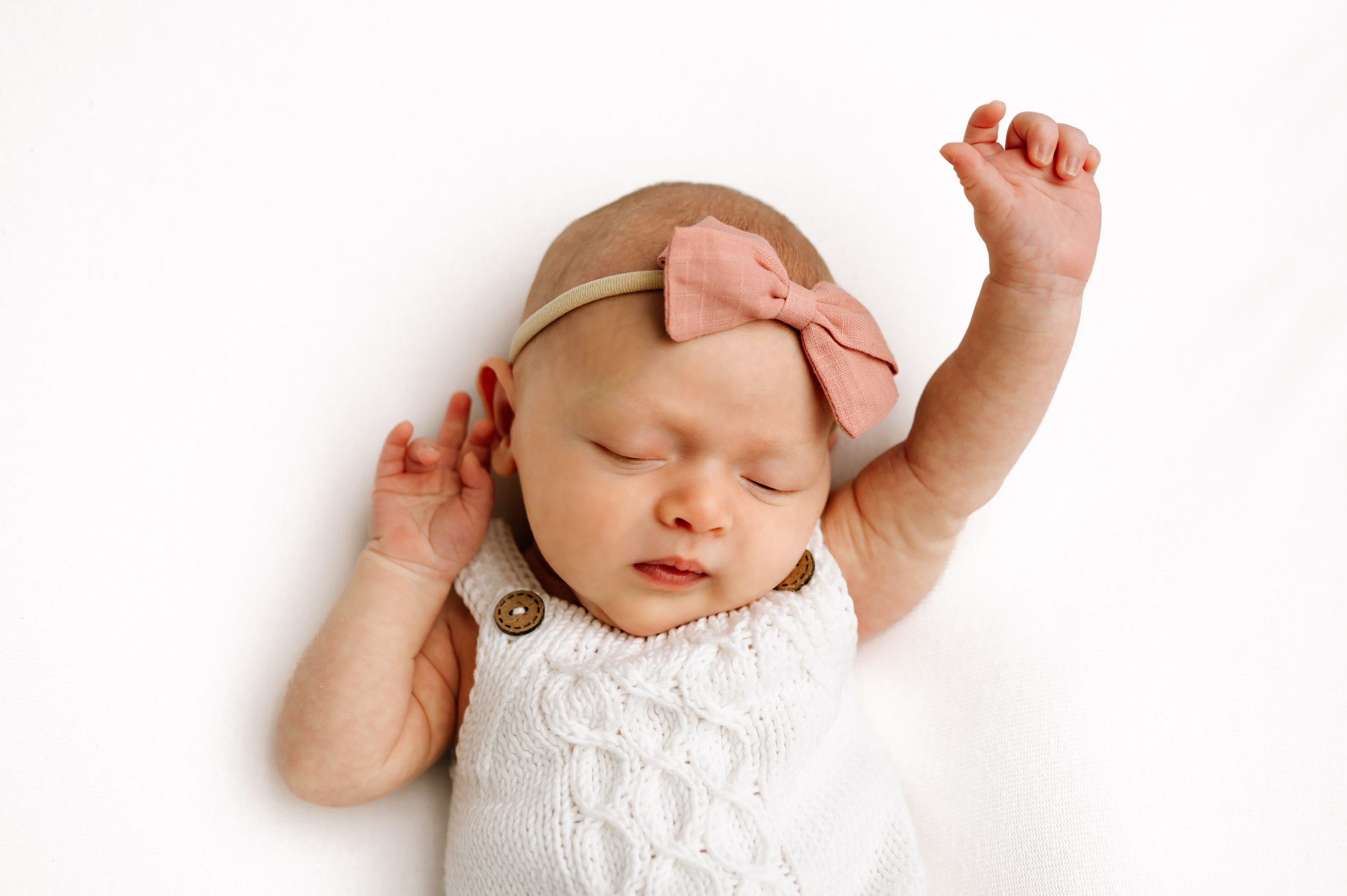 a baby girl wearing a white knit romper and a pink headband laying on a white backdrop and stretching her arms as she wakes up from a nap during a newborn photoshoot