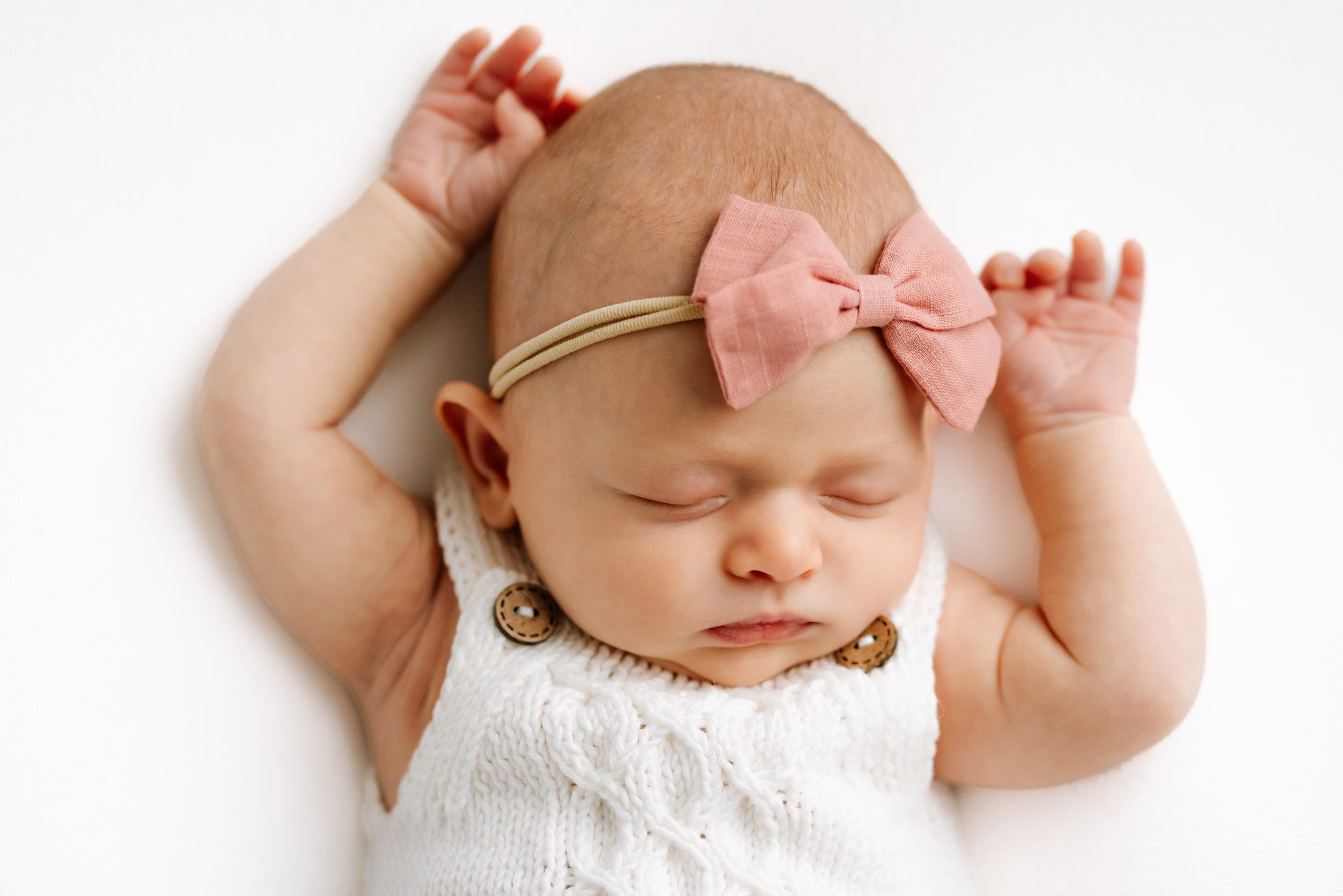 a baby girl wearing a white knit romper and pink headband laying on a white backdrop and sleeping peacefully with her arms resting up next to her head during a newborn photo session