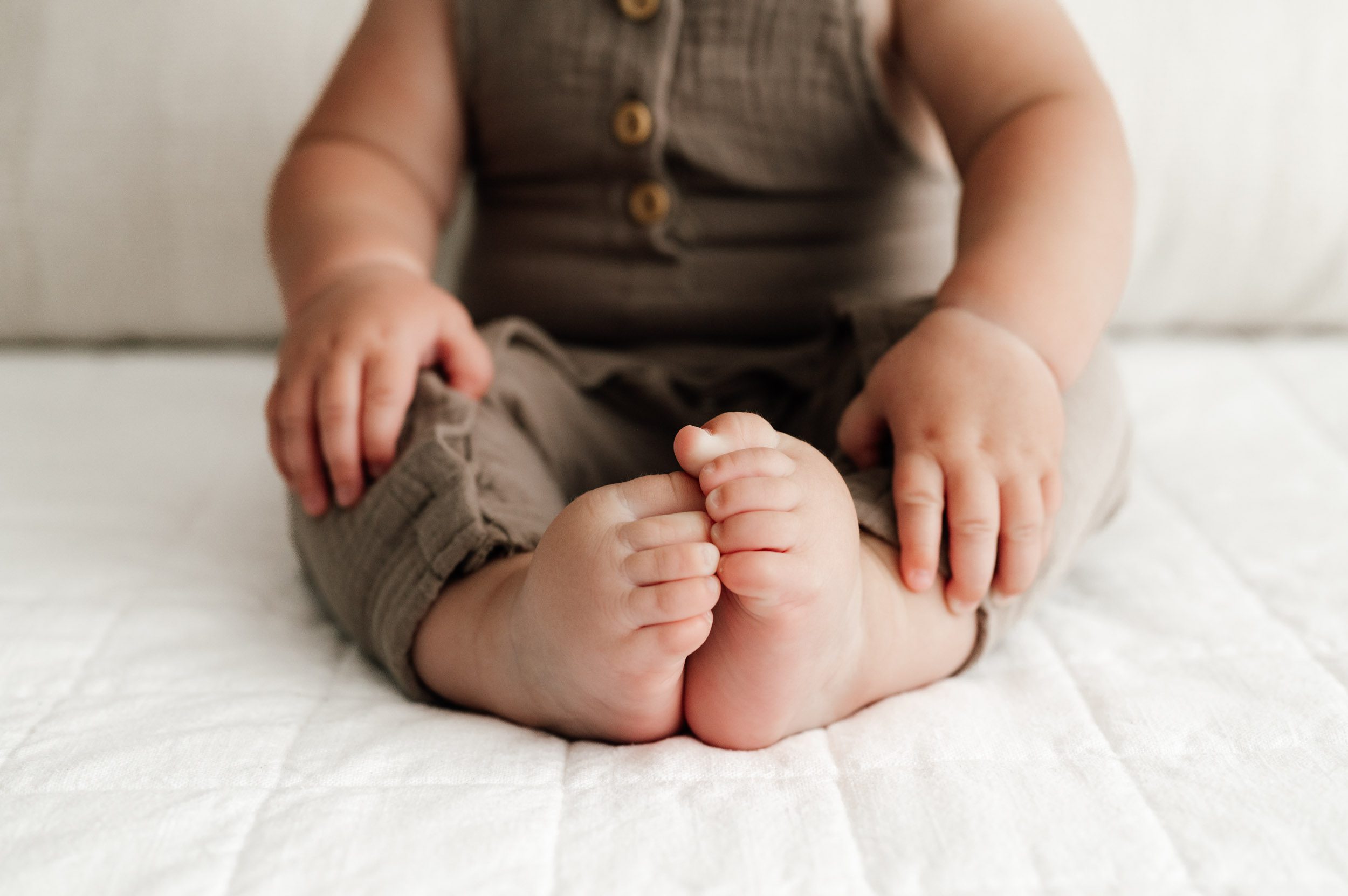 a close up picture of a baby boy's feet and hands as he sits on a bed during a baby milestone photo session