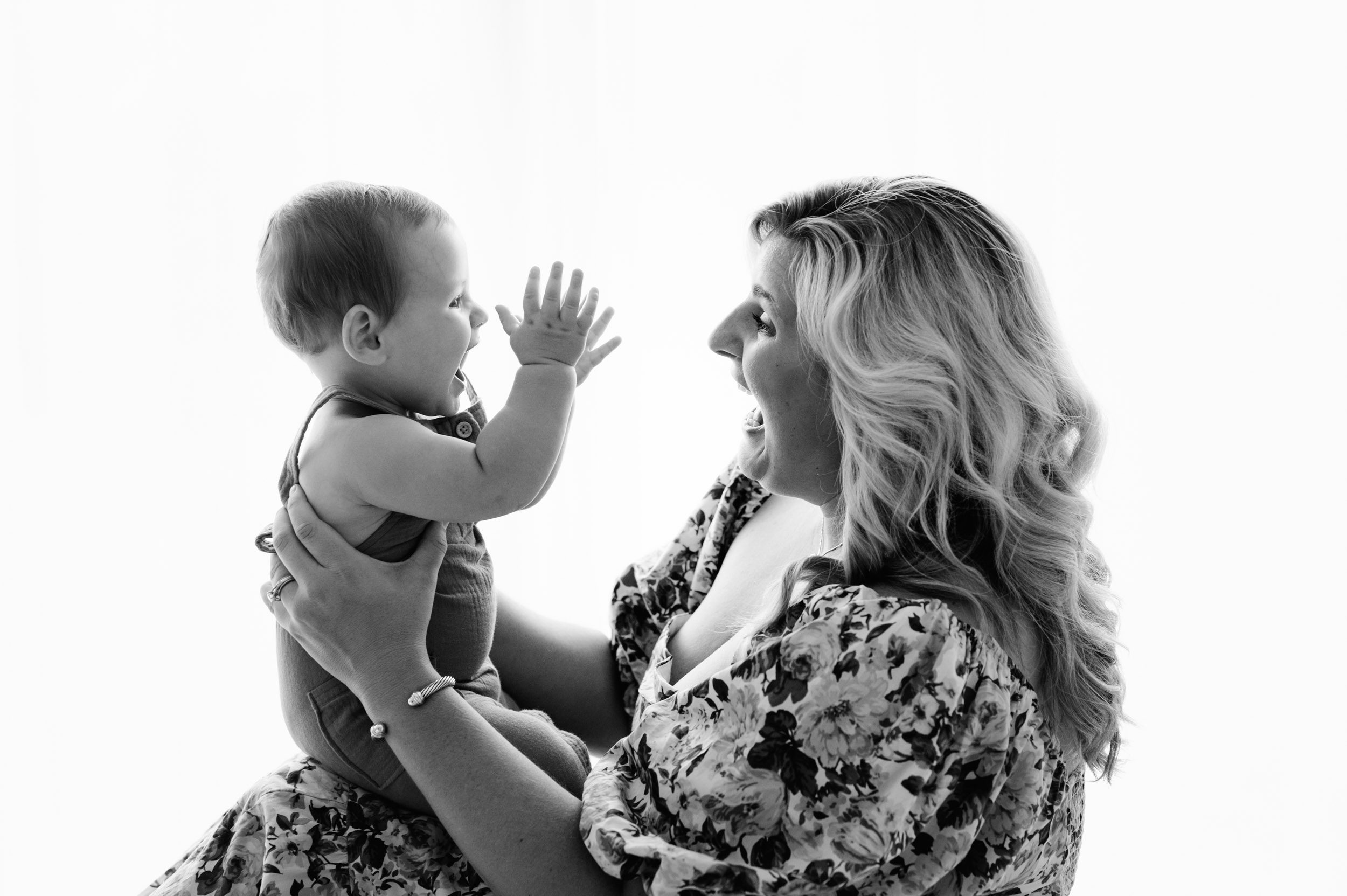 a black and white backlit photo of a baby boy sitting on his mom's lap and clapping as they both laugh during a baby's first year milestone photoshoot