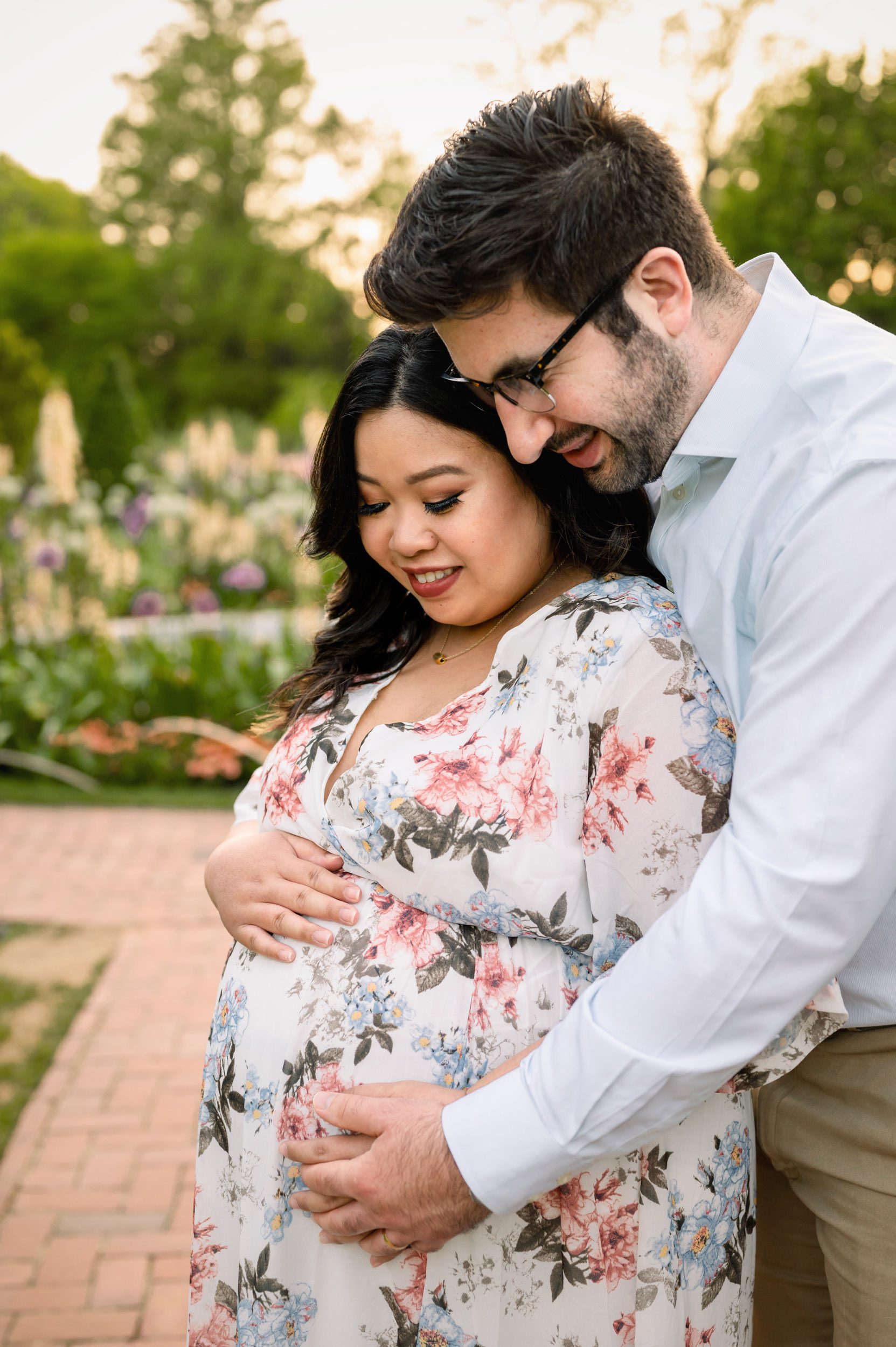 an expecting couple standing on a brick pathway with colorful flowers blooming all around them as dad hugs mom from behind and they both smile down at her belly during a maternity photo session