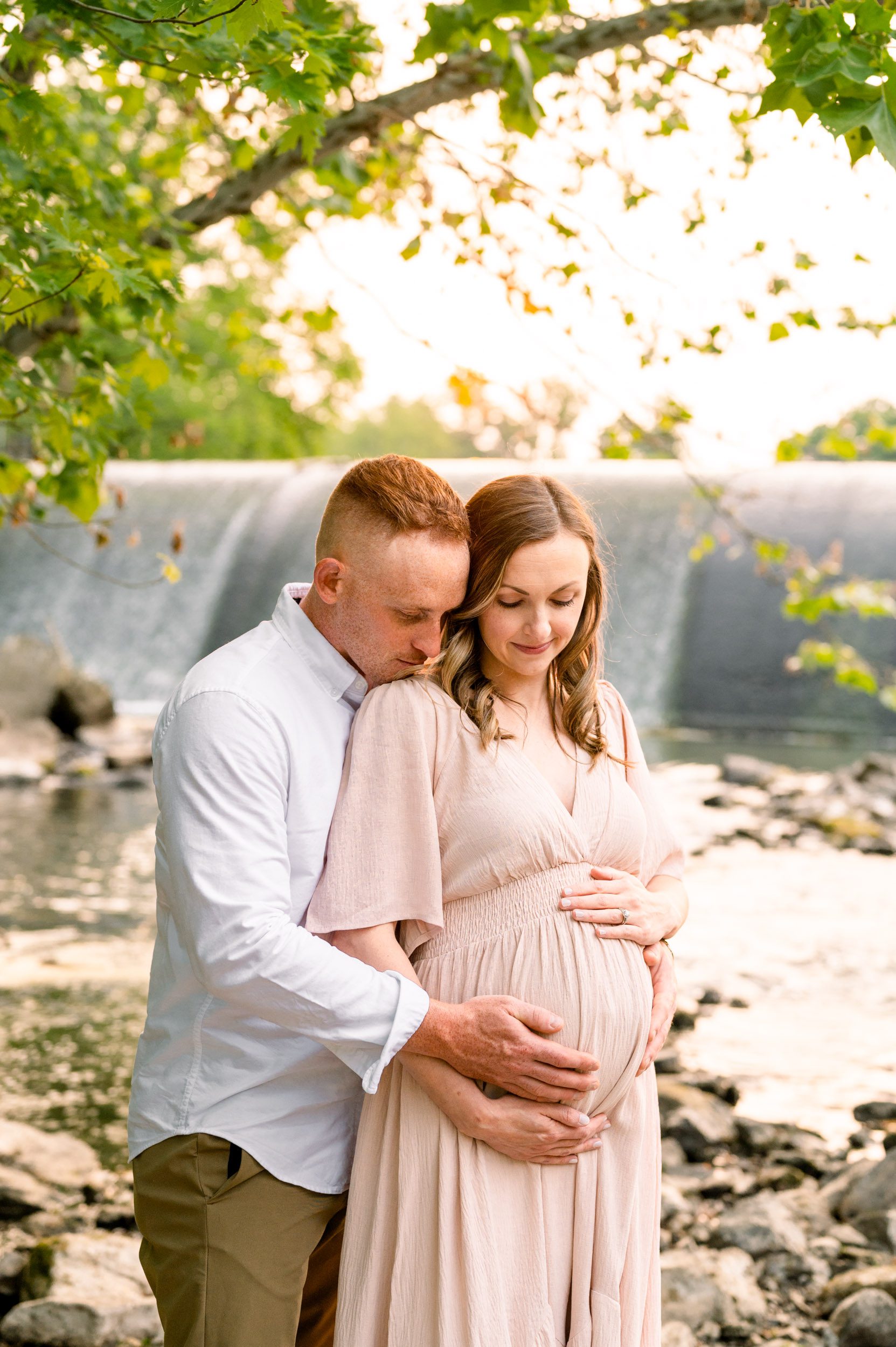 expecting parents standing in front of a waterfall as dad hugs mom from behind and they both smile down at her belly during a maternity photo session