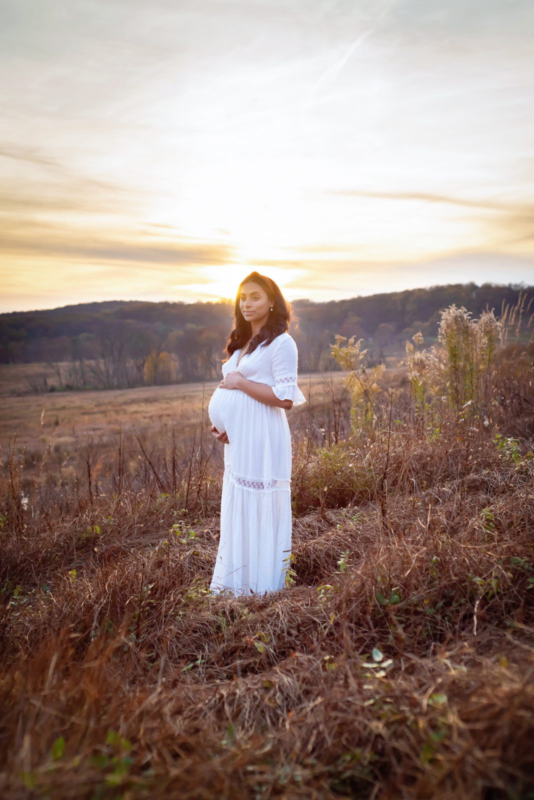 an expecting mother wearing a white dress standing in a field of golden grasses with the sunset shining in the background during a maternity photo session