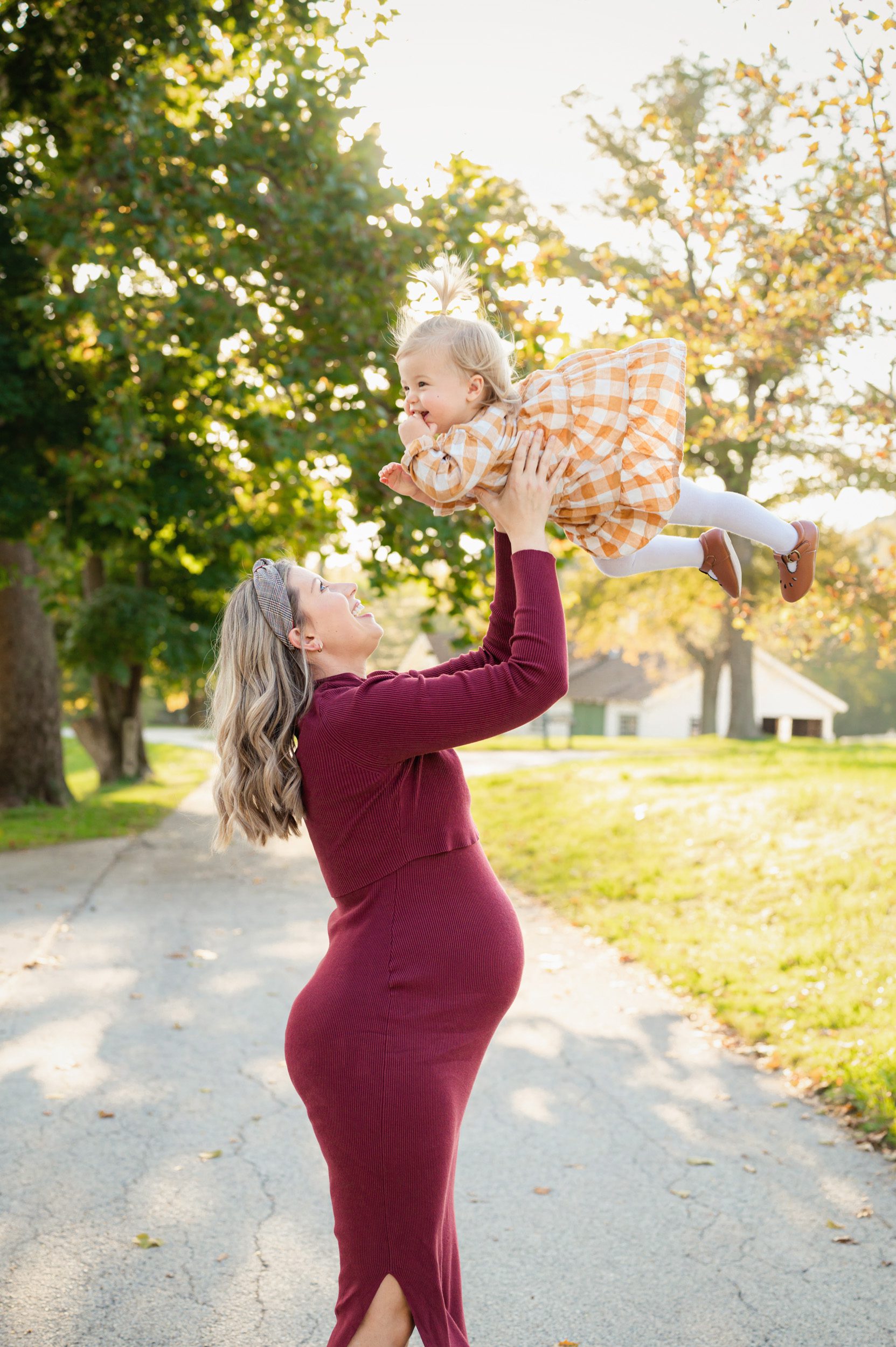 an expecting mom wearing a burgandy dress and lifting her young daughter up in the air during a maternity photoshoot