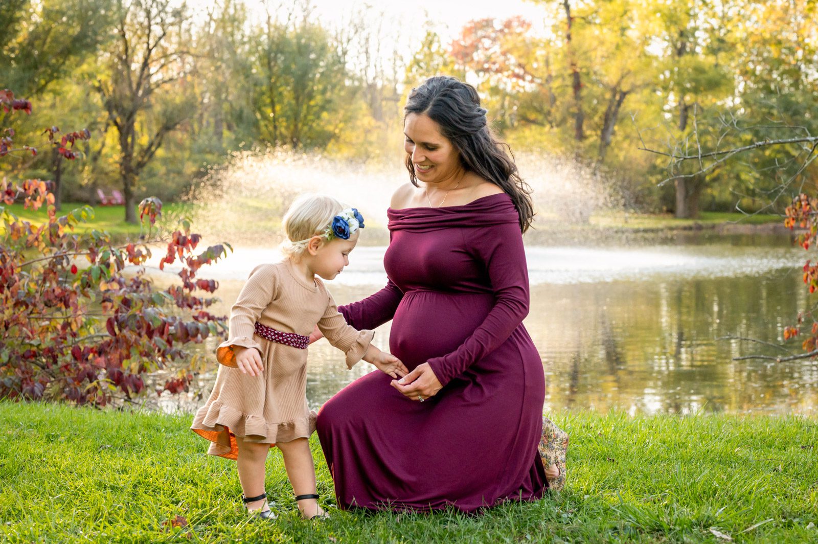 an expecting mom kneeling down in front of a pond while her young daughter reaches out to gently touch her belly during a maternity photo session