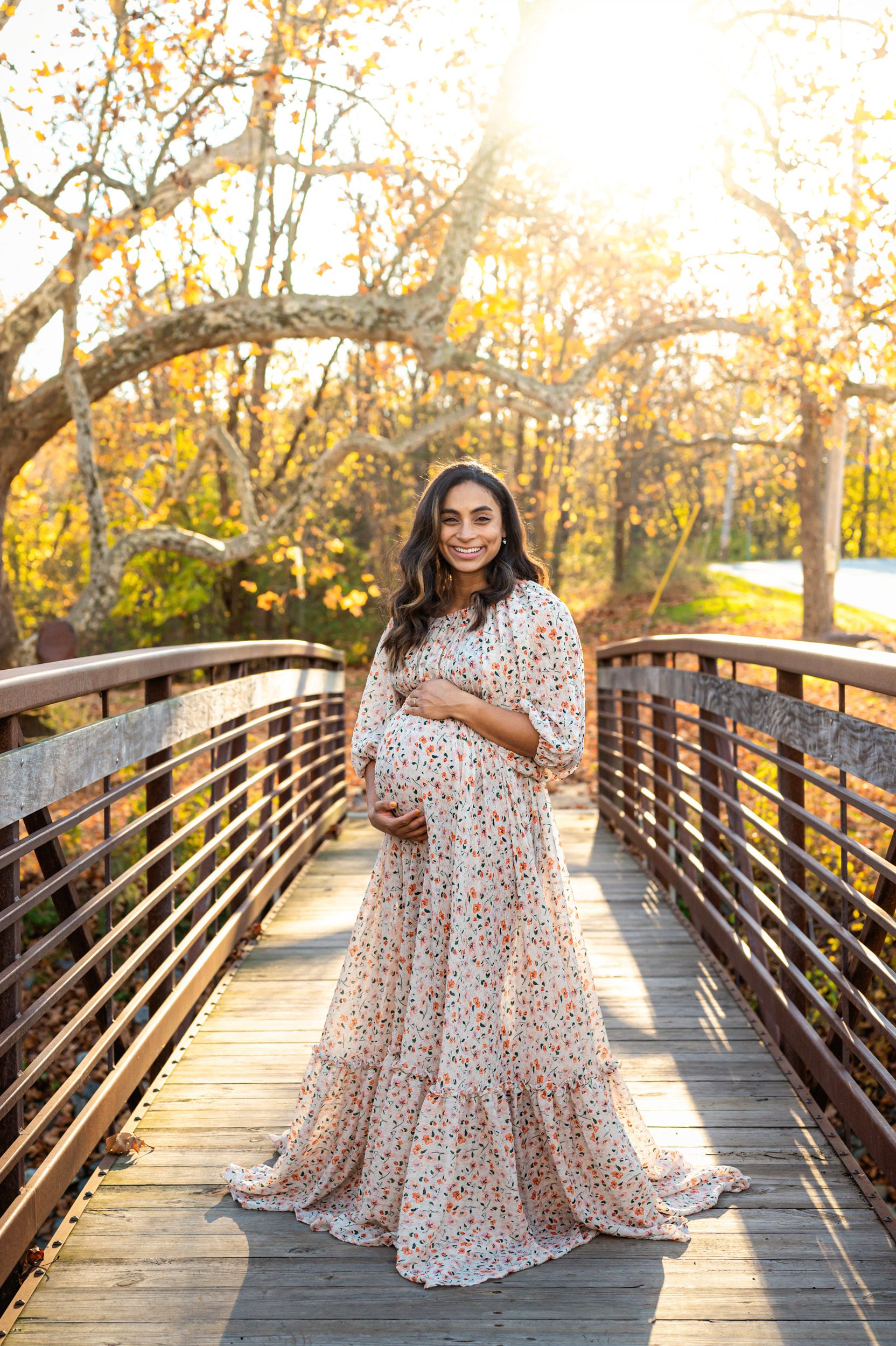 an expecting mother wearing a floral dress standing on a bridge and smiling at the camera with the sunset shining through the colorful fall leaves in the background during a maternity photoshoot