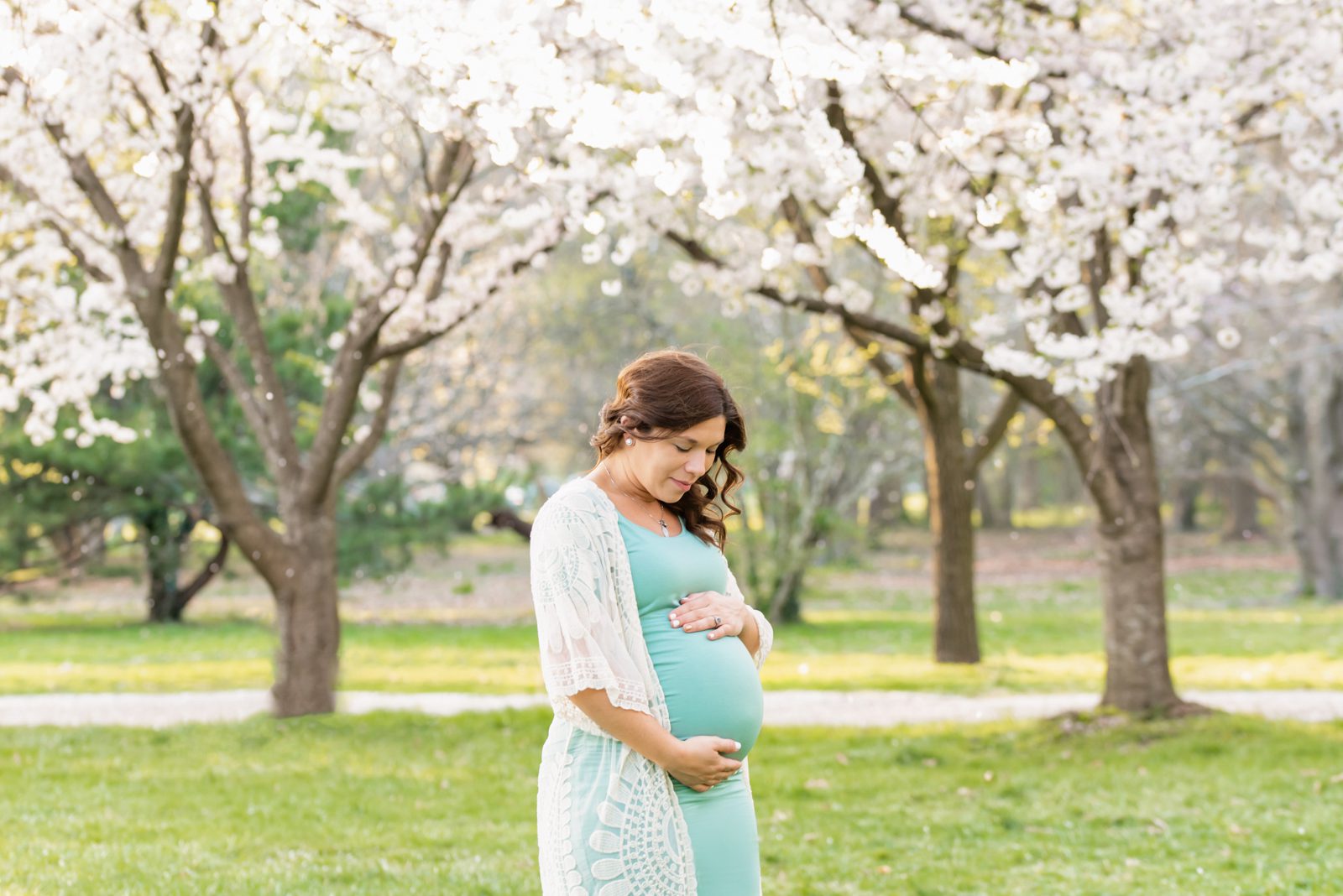an expecting mom standing in the grass and cradling her belly with white cherry blossom trees in bloom behind her during a maternity photo session