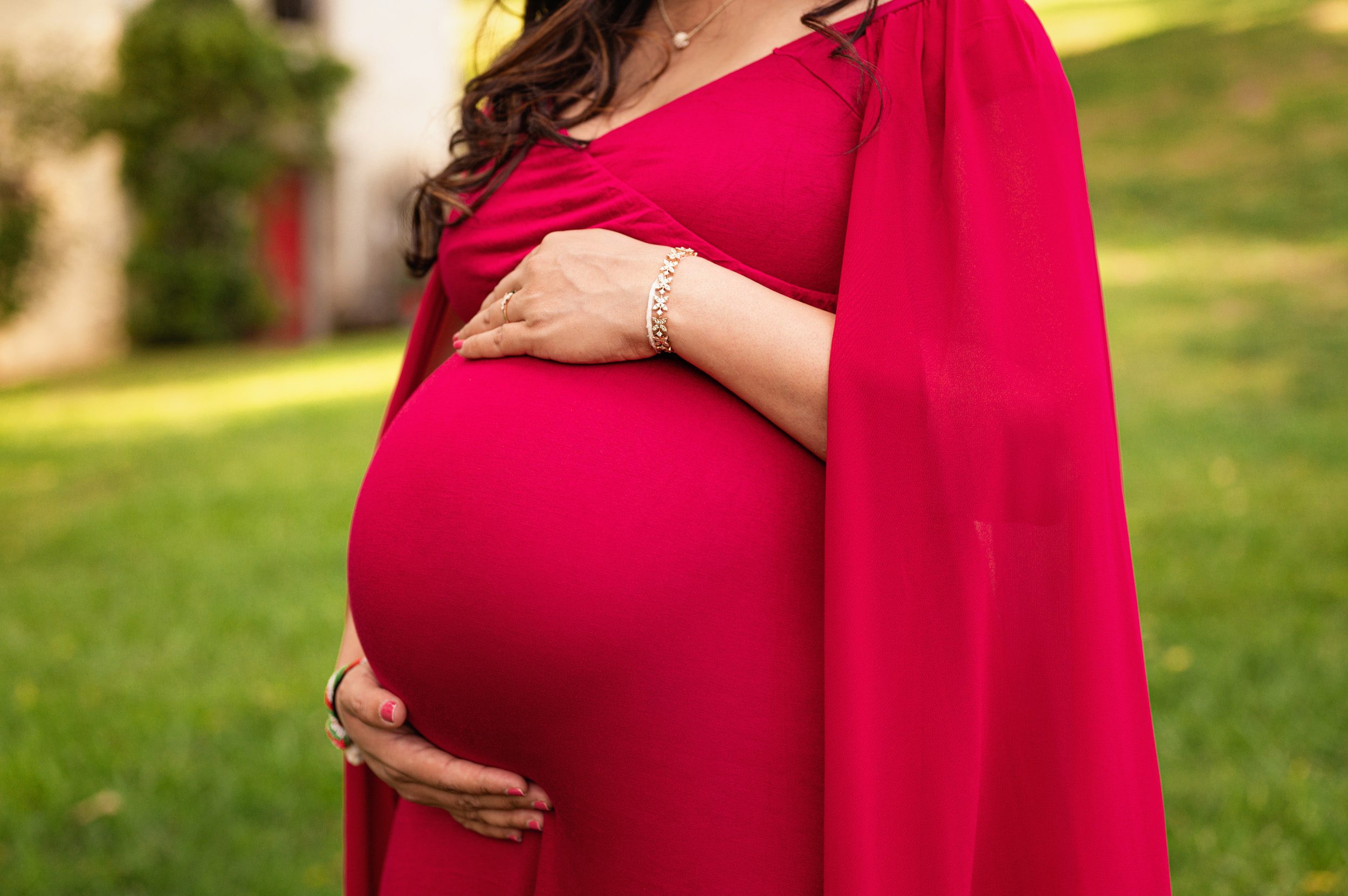 a close up picture of an expecting mom wearing a red dress cradling her belly in her hands 