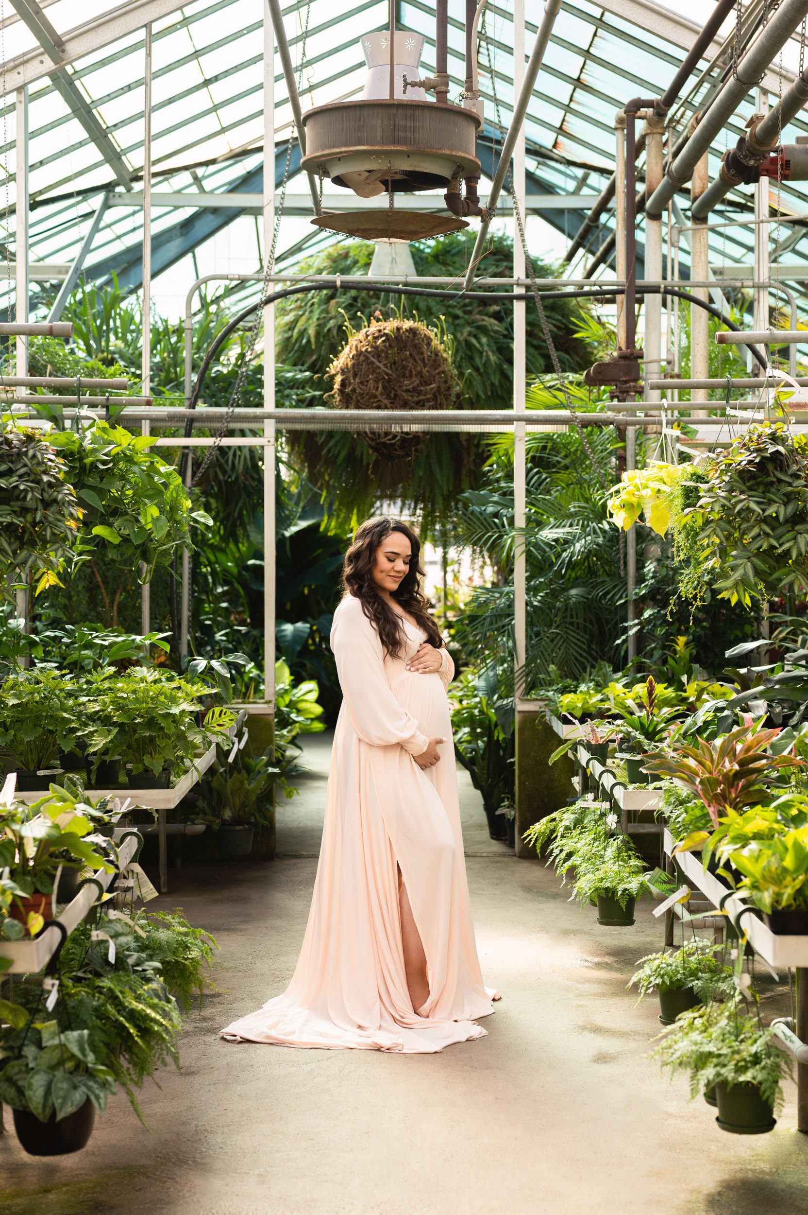 an expecting mom wearing a light pink dress standing in a greenhouse and smiling down at her belly during a maternity phootshoot