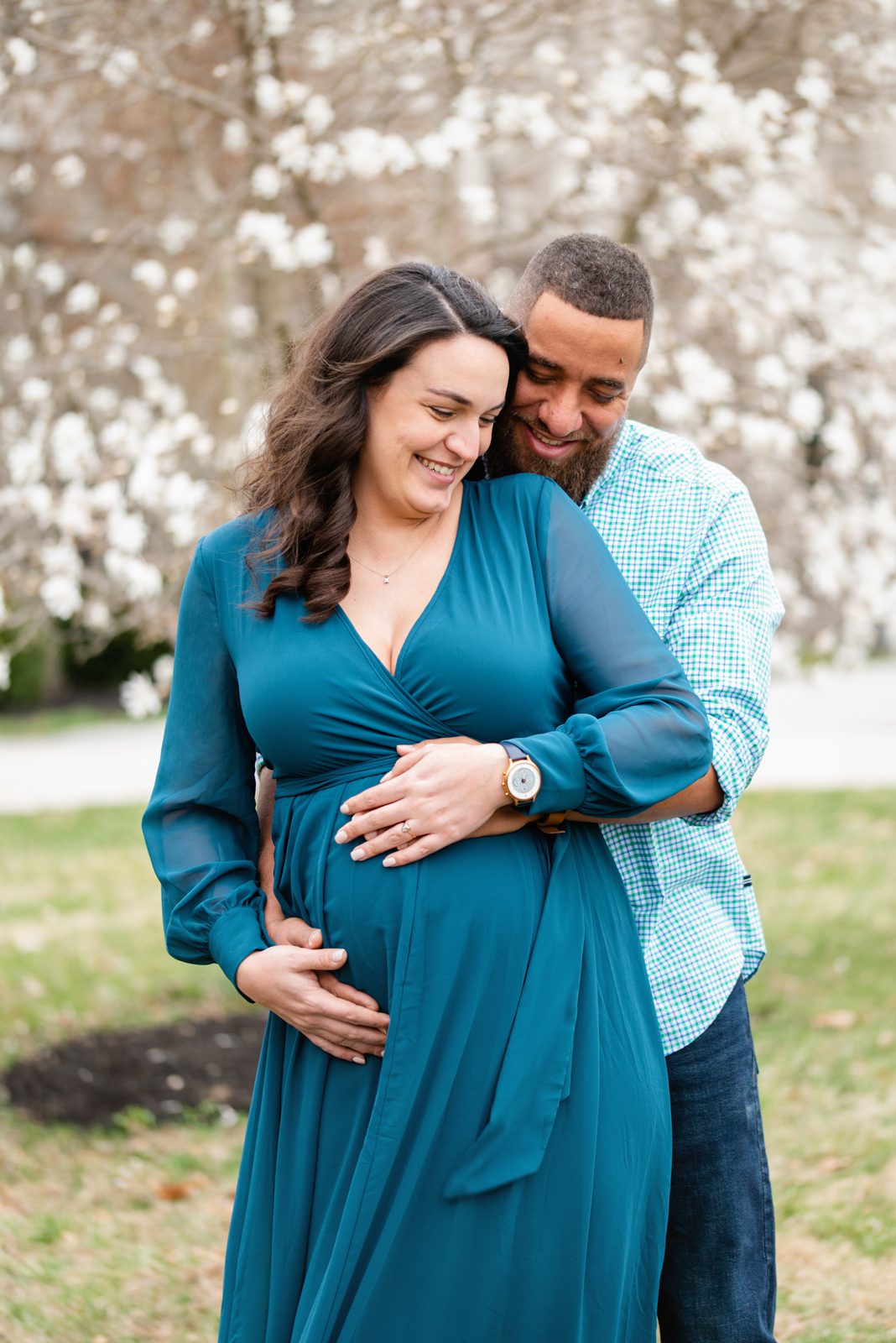 an expecting couple standing in front of a tree full of white flowers and smiling as dad hugs mom from behind during a maternity photo session