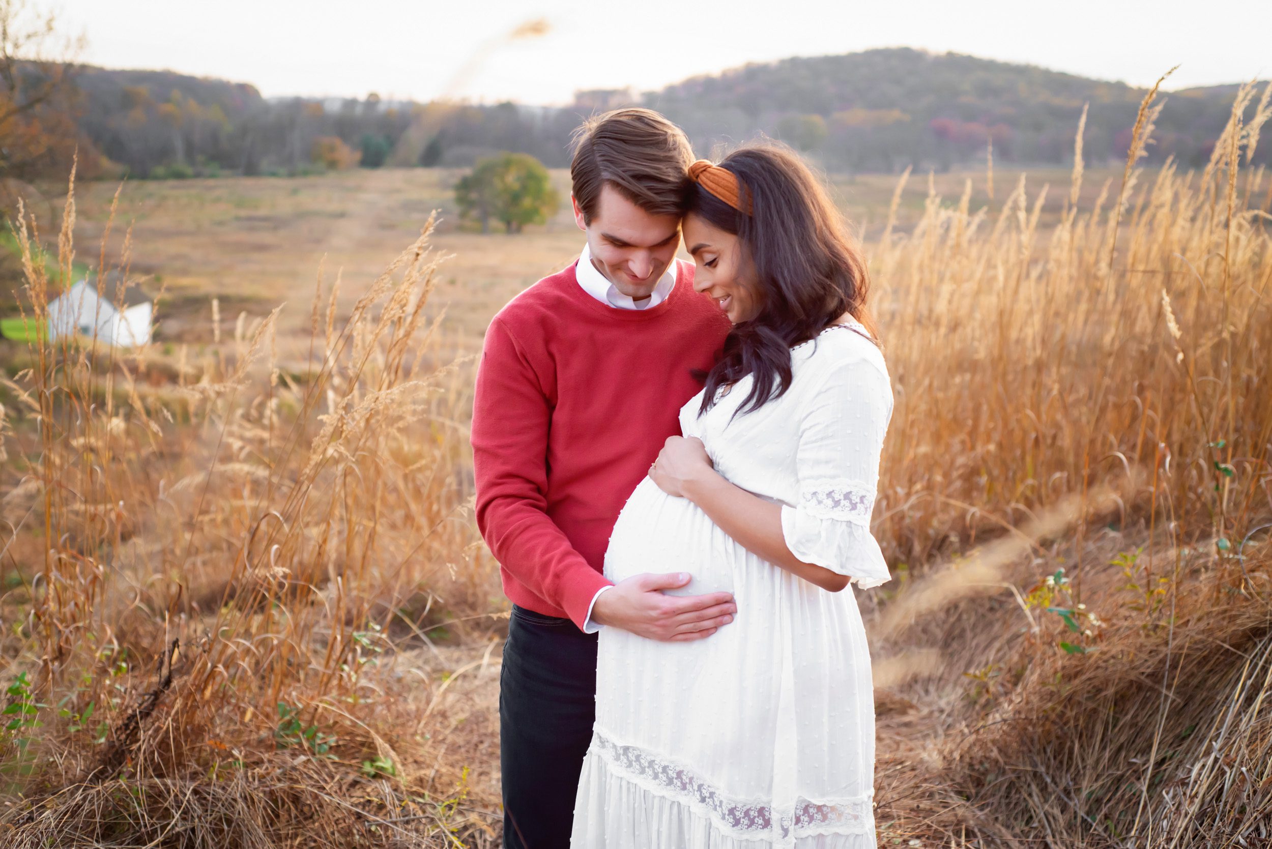an expecting couple standing in a field of golden grasses and resting their heads together as they both smile down at mom's belly
