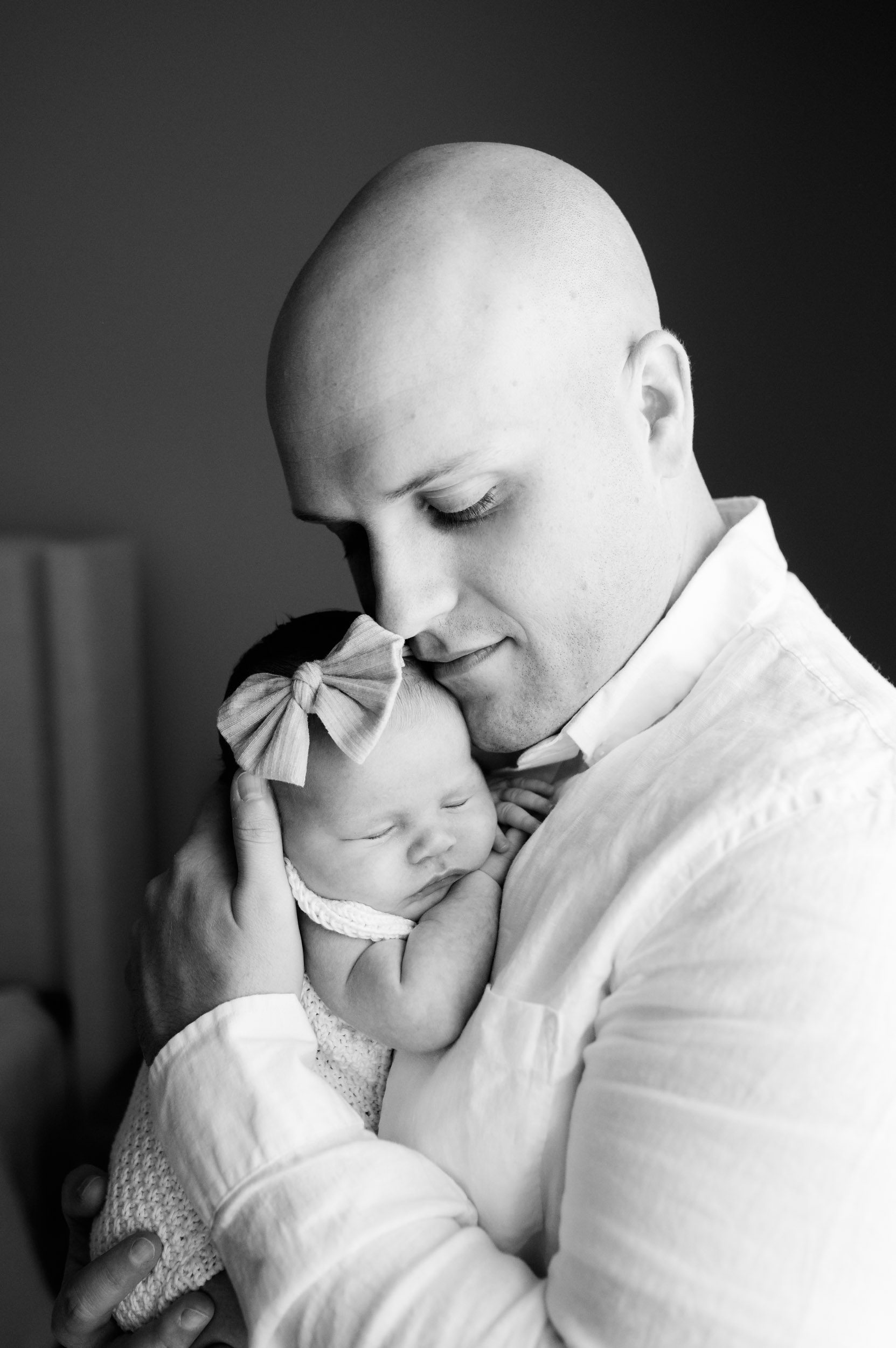 a black and white picture of a new father holding his baby girl snuggled up against his chest as she sleeps during an in home newborn photo session