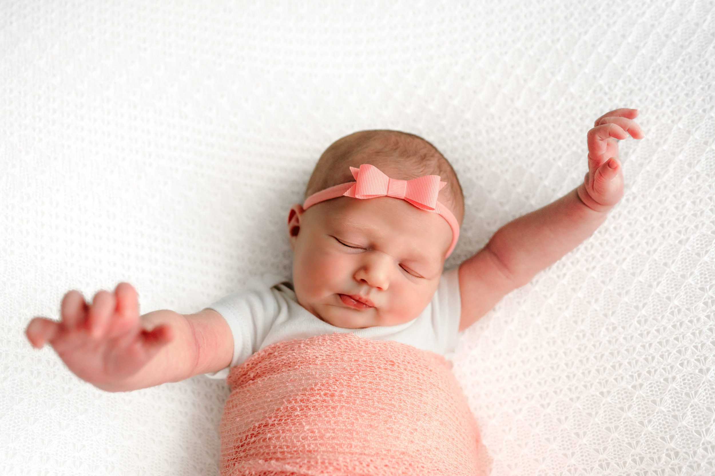 a baby girl wearing a pink headband wrapped in a pink swaddle blanket laying on a white textured backdrop stretching her arms as she sleeps during an in home newborn photoshoot