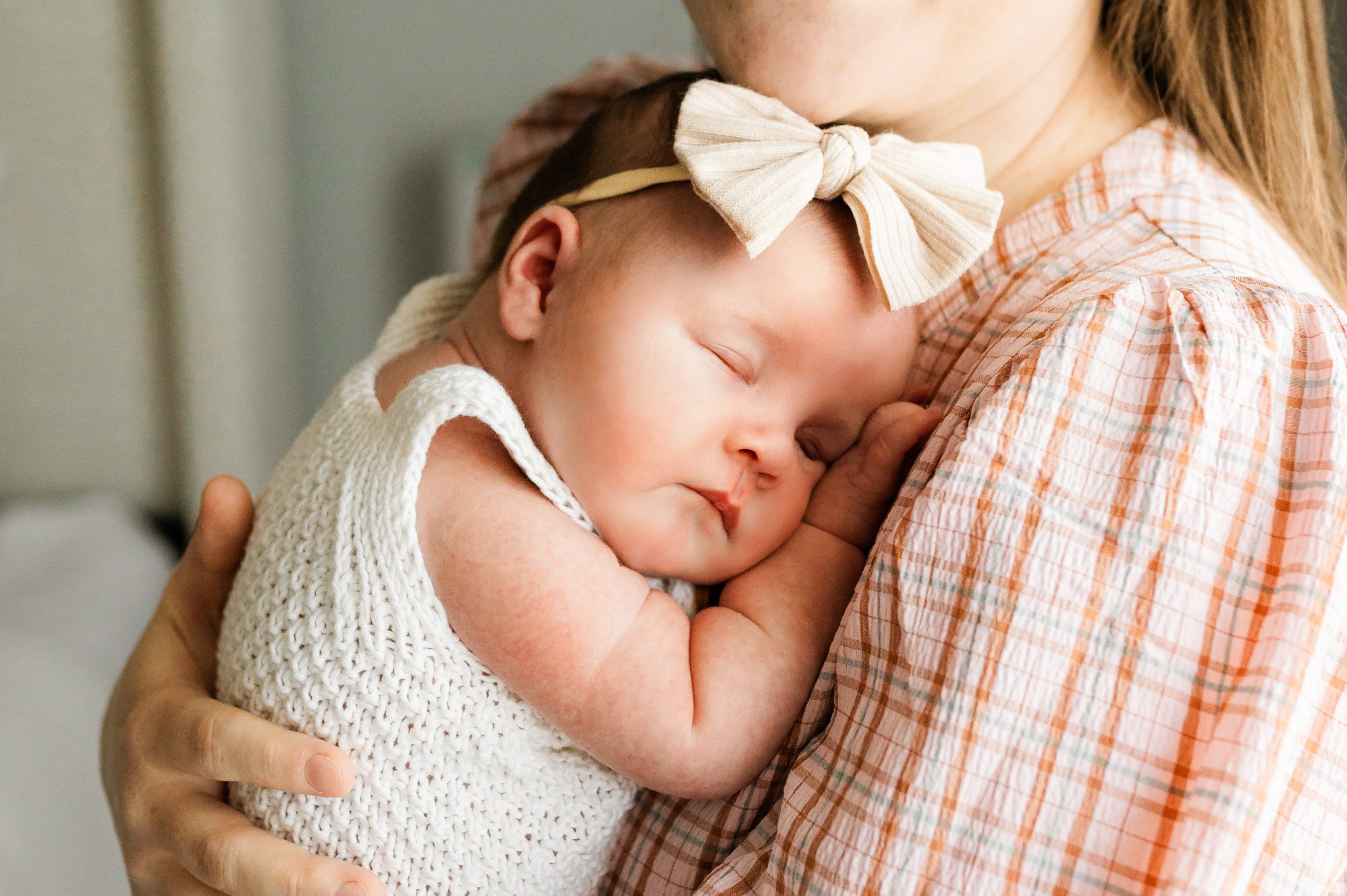 a close up picture of a baby girl wearing a white textured knit romper sleeping peacefully snuggled up against her mom's chest during a Collegeville in home newborn photo session