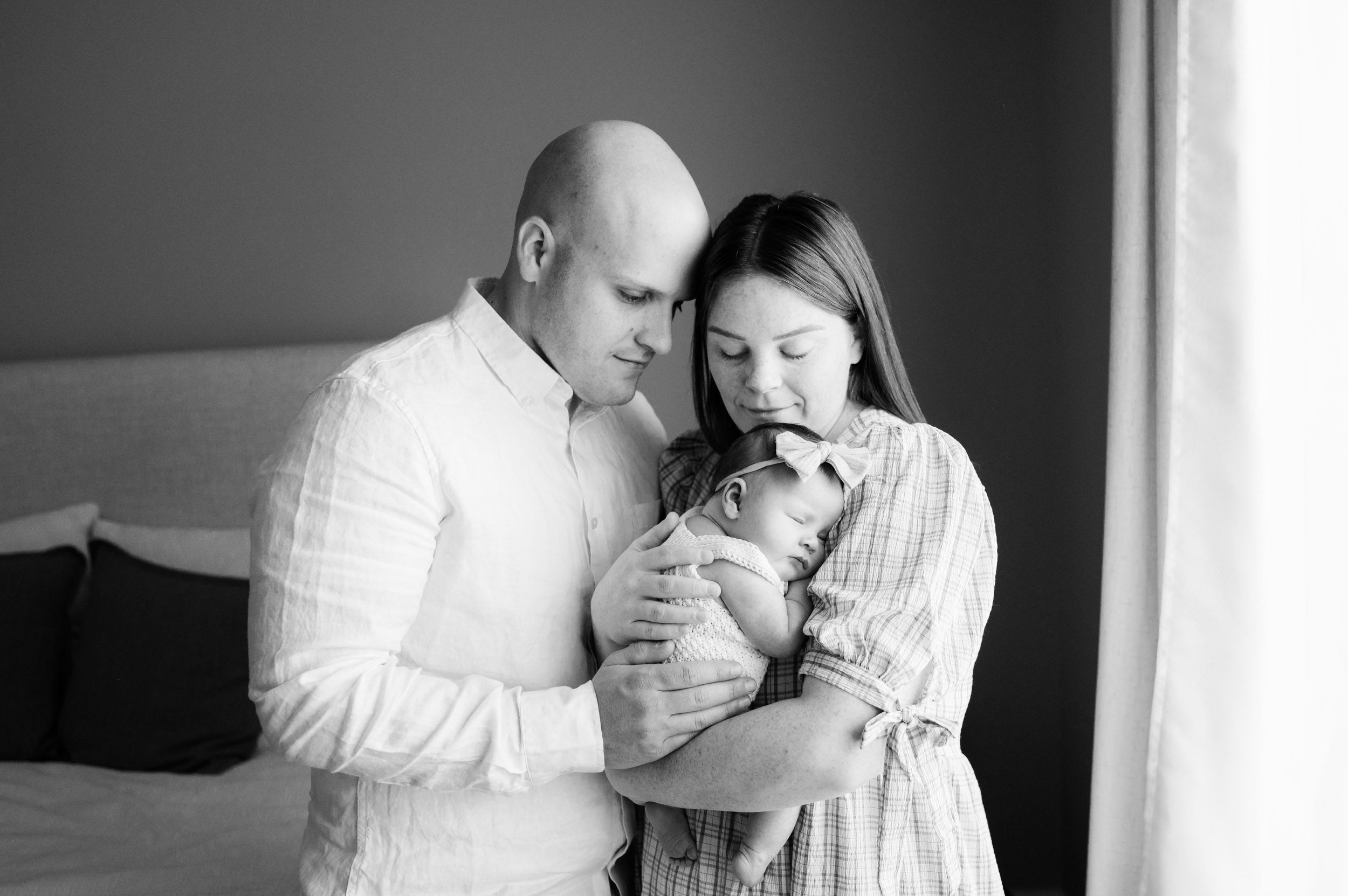 a black and white picture of a baby girl sleeping peacefully snuggled up against her mom's chest as mom and dad rest their foreheads together and smile down at her during a Collegeville in home newborn photoshoot
