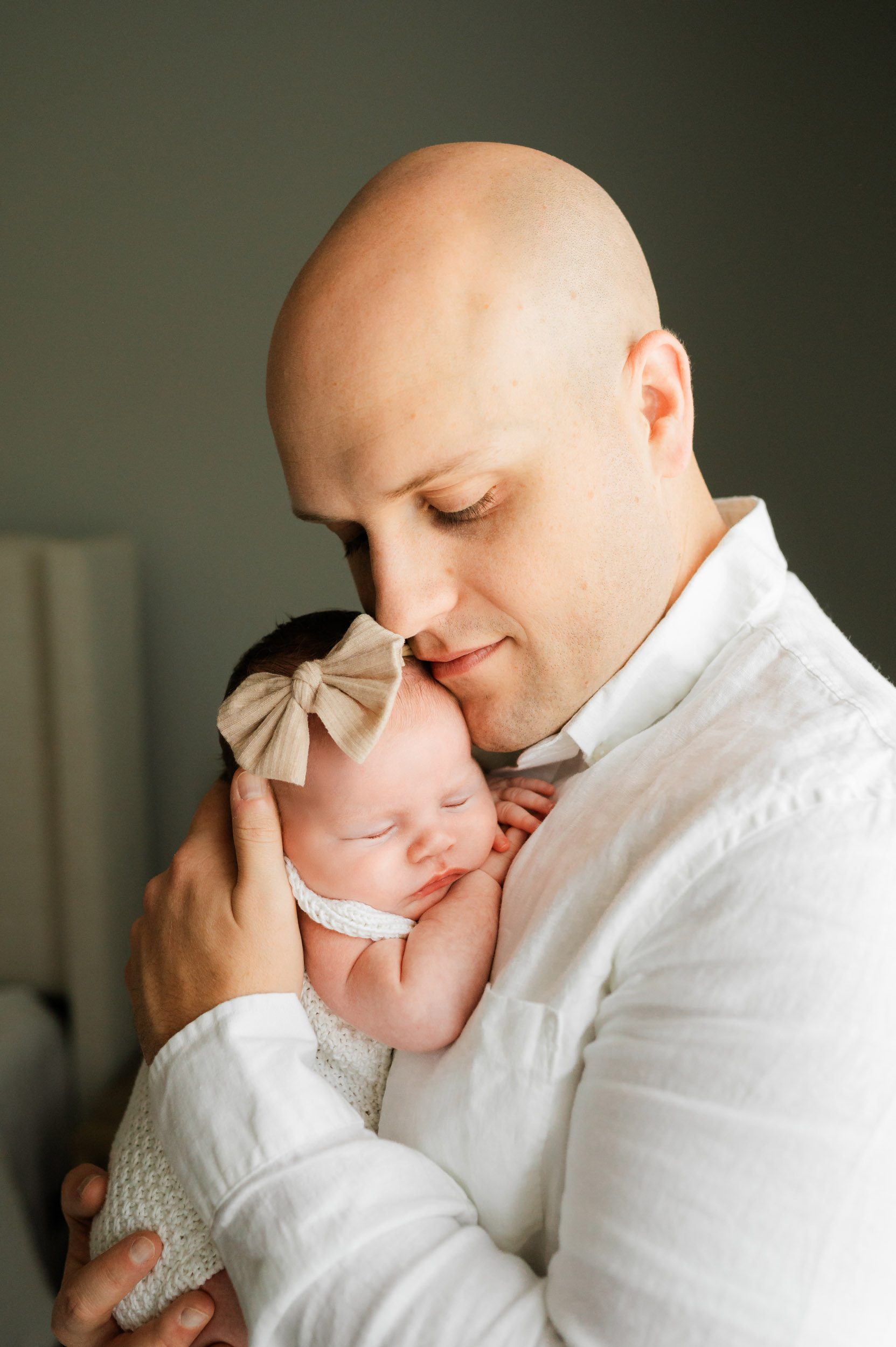 a baby girl wearing a textured white romper and beige bow headband sleeping peacefully snuggled up against her dad's chest as he smiles down at her during a Collegeville newborn photo session