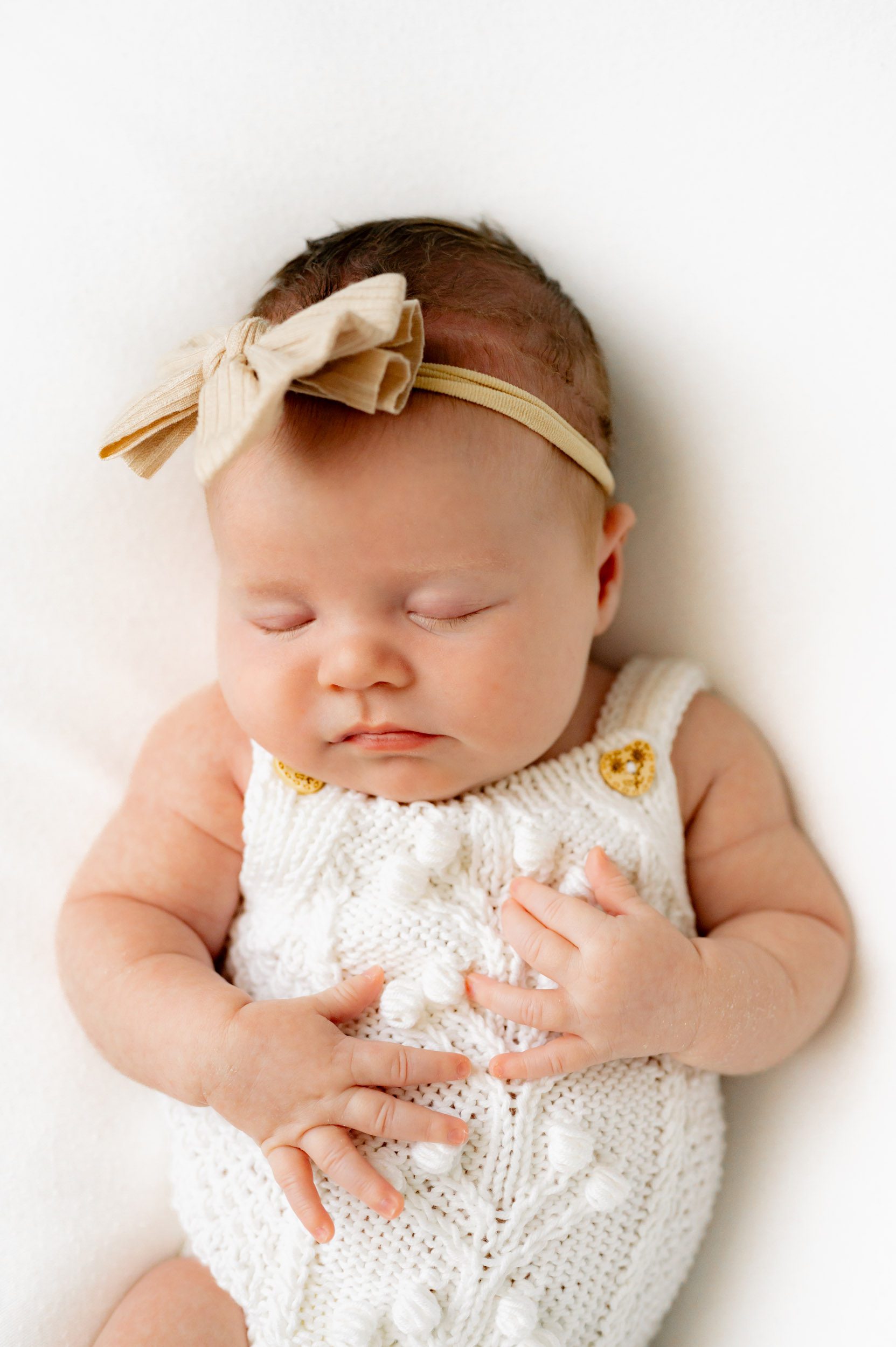 a baby girl wearing a white textured knit romper and beige headband laying on a white backdrop and sleeping peacefully during a newborn photoshoot
