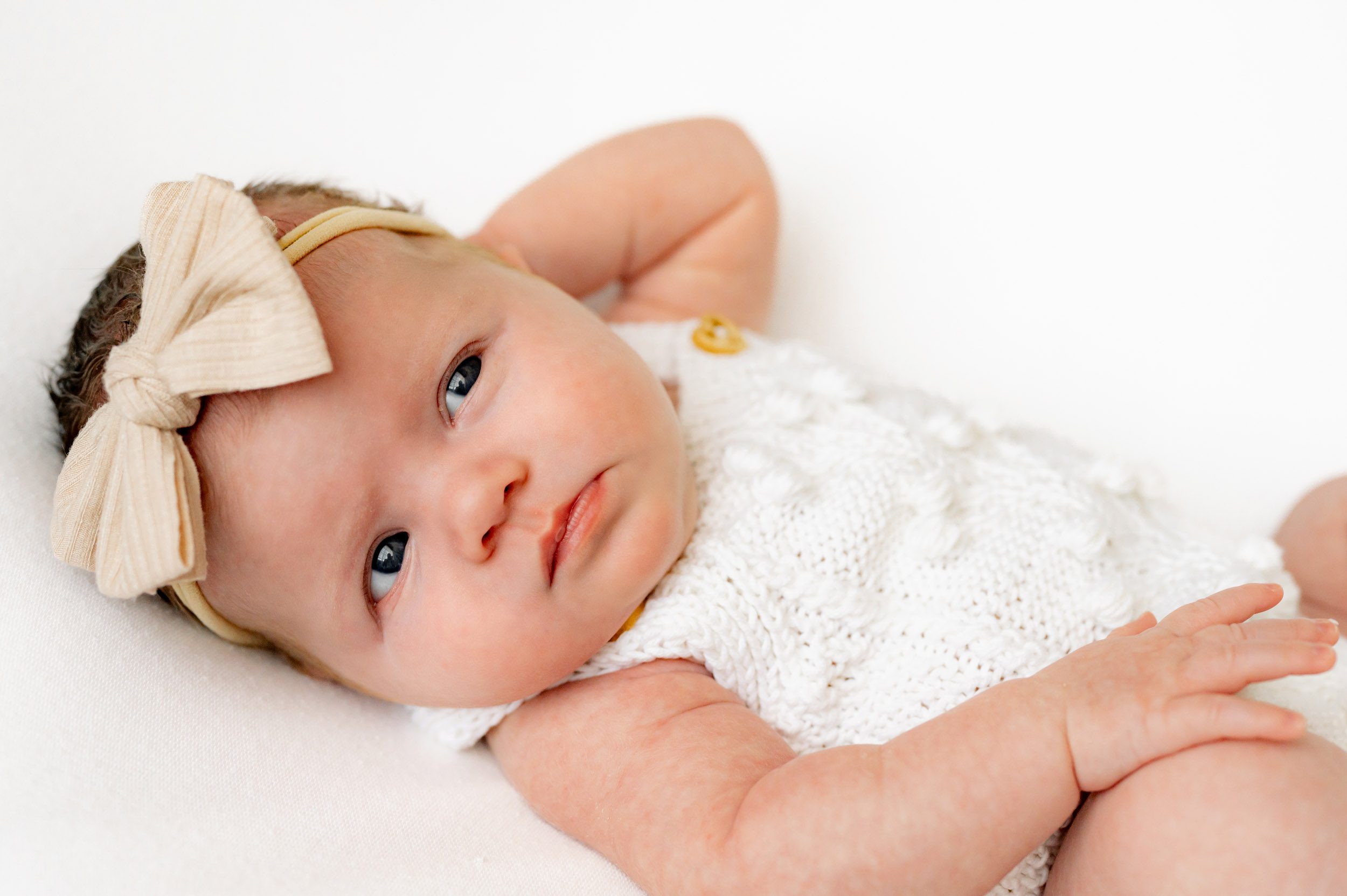 a baby girl wearing a white textured knit romper and beige bow headband laying on a white backdrop and touching her knee as she looks off toward the window with catch lights in her eyes