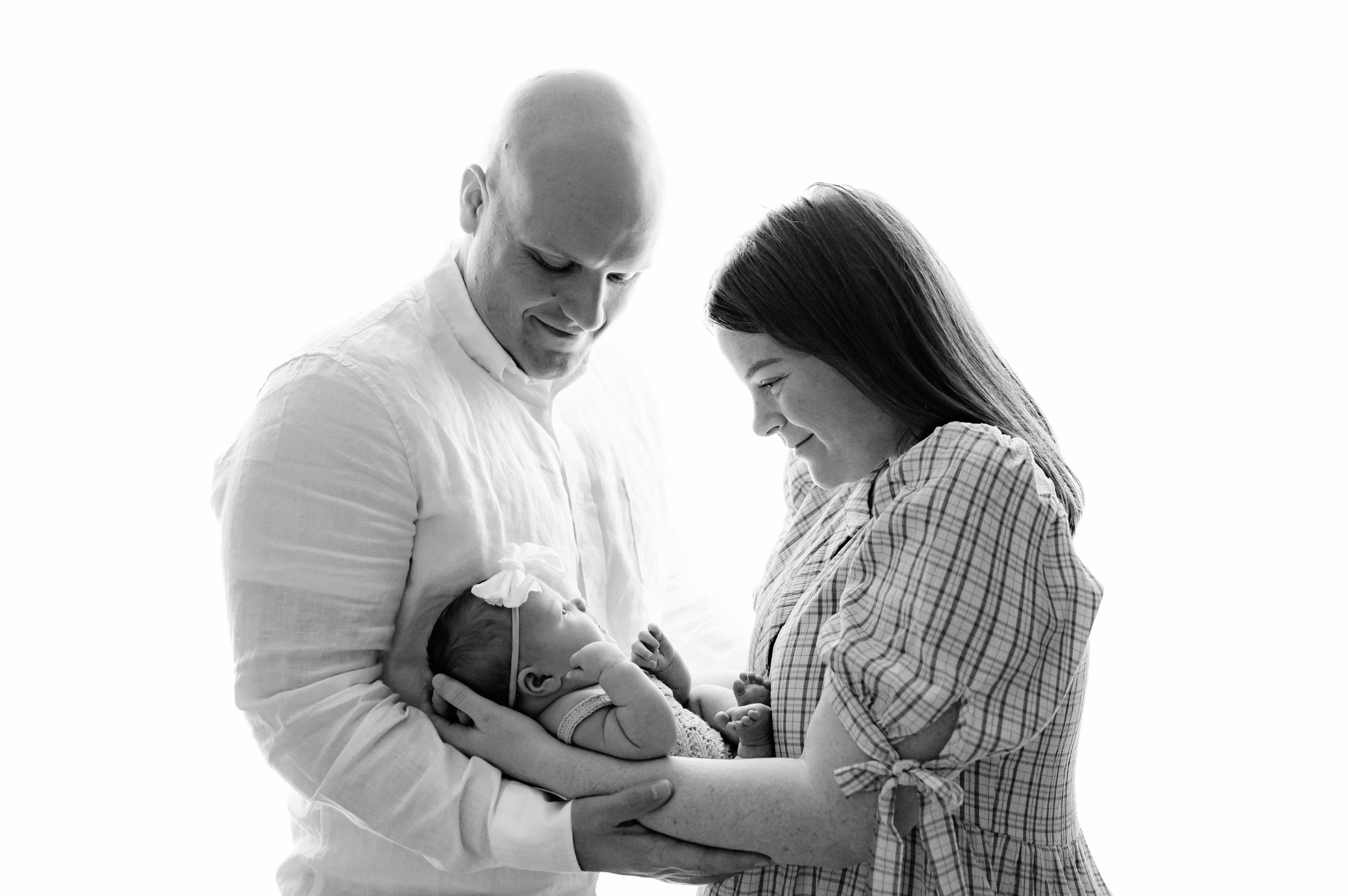 a black and white backlit picture of new parents holding their baby girl in their arms and smiling down at her during a newborn photoshoot