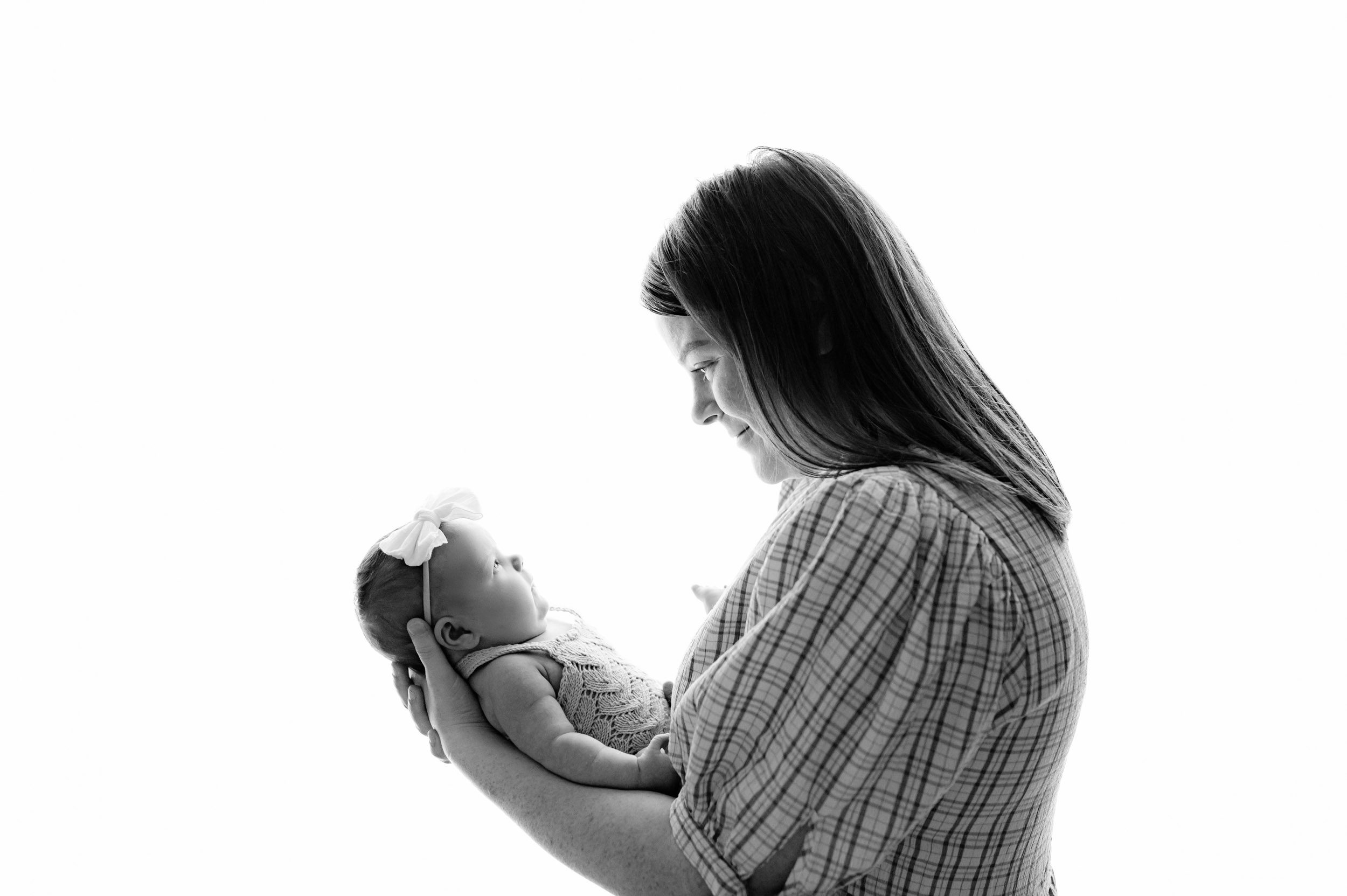a black and white backlit photo of a new mom holding her baby girl in her arms and smiling down at her during an in home newborn photoshoot