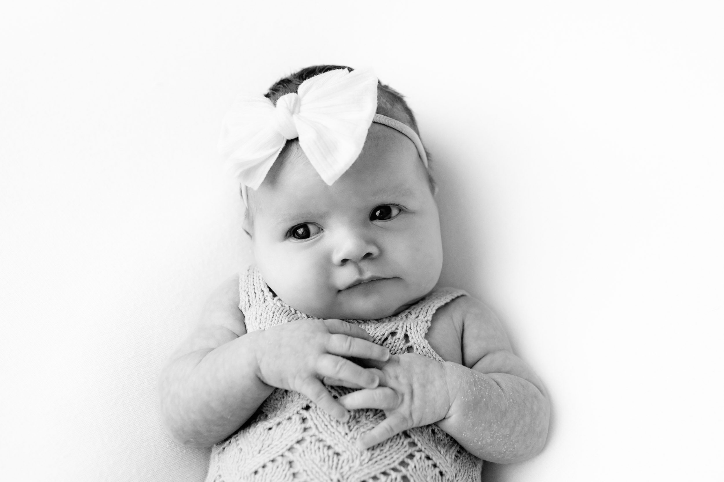 a black and white picture of a baby girl wearing a textured knit romper and a white bow headband laying on a white backdrop and looking off toward the side with wide, curious eyes during a newborn photo session