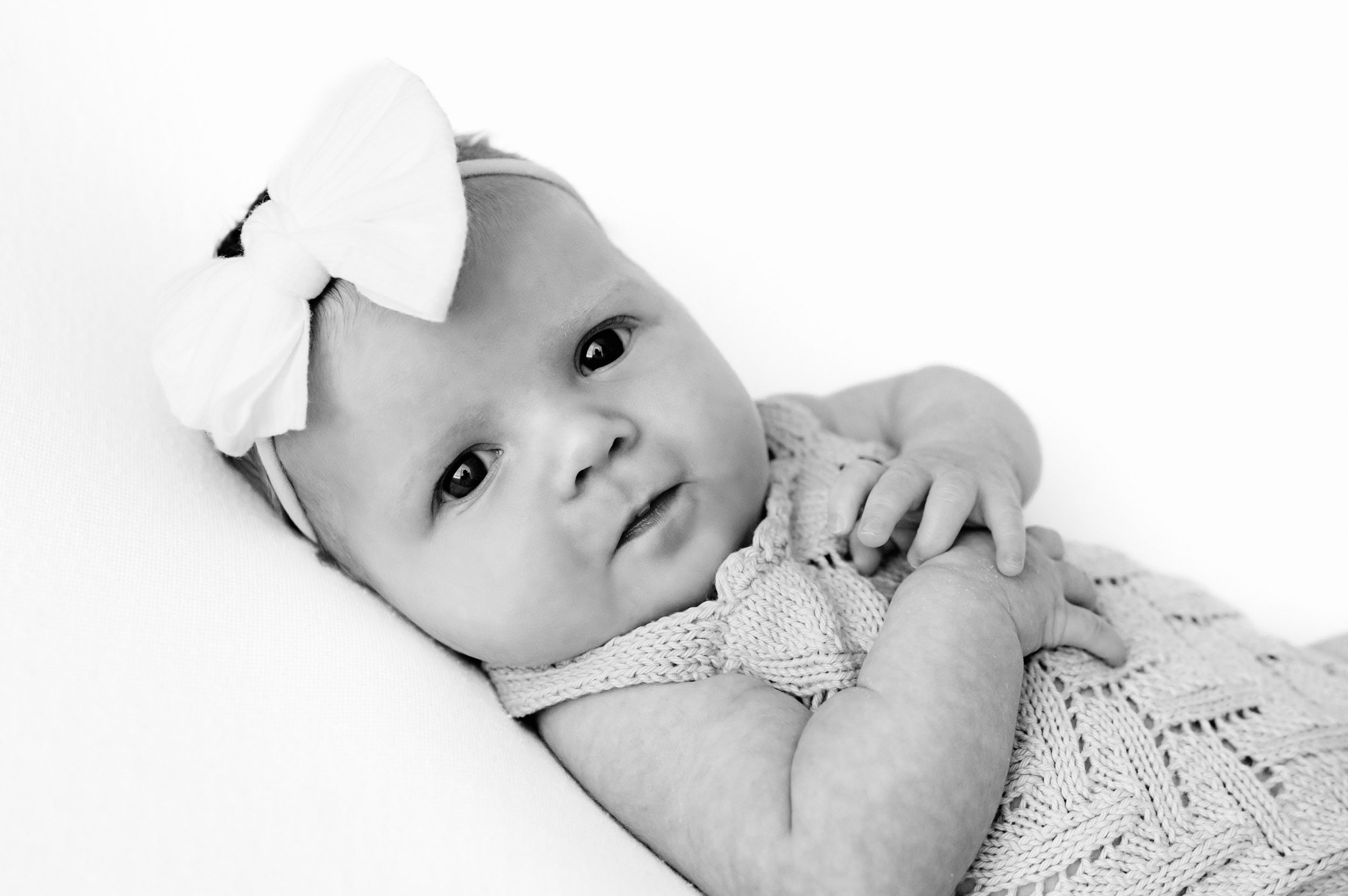 a black and white picture of a baby girl wearing a textured knit romper and a white bow headband laying on a white backdrop and looking directly at the camera with a hint of a smile on her face during an in home newborn photoshoot
