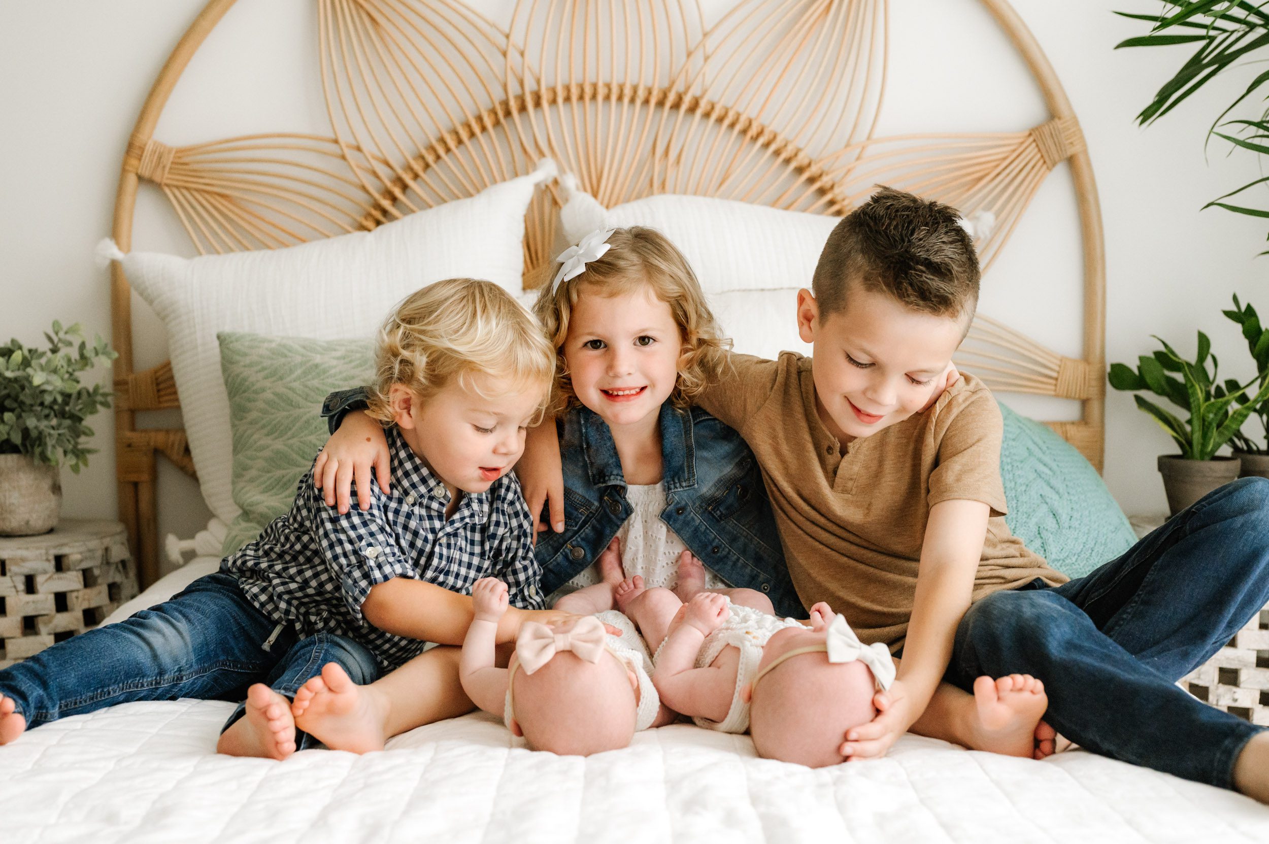 twin baby girls laying on a bed while their two older brothers smile down at them and touch them with their hands and their big sister smiles right at the camera during a newborn photoshoot