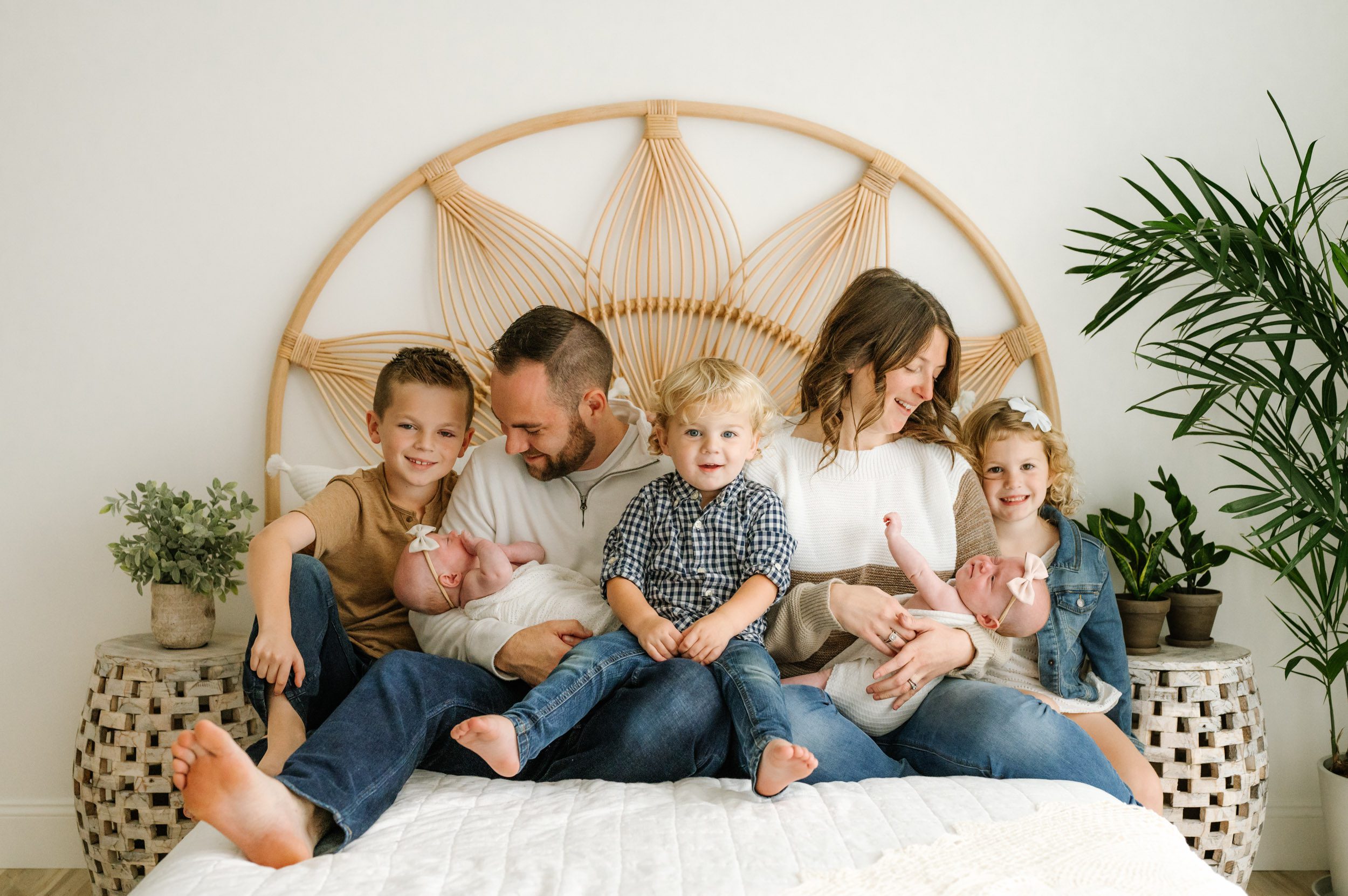 a family of seven sitting on a bed with mom and dad holding their twin baby girls in their arms and smiling down at them and their three older kids smile at the camera during a newborn photo session