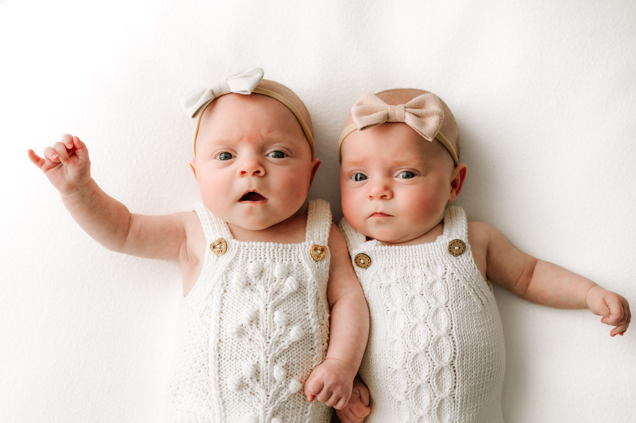 twin baby girls wearing white knit rompers and laying on a white backdrop looking directly at the camera during a newborn photoshoot