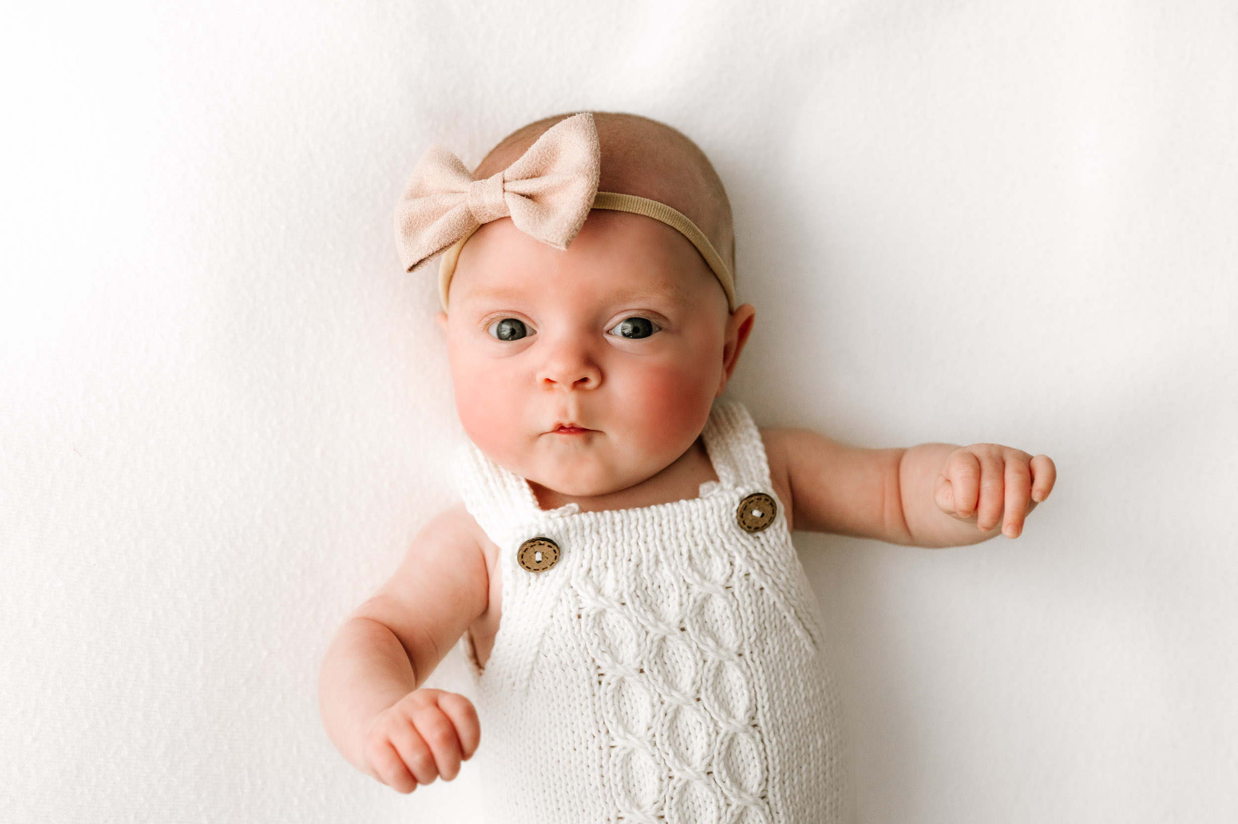 a baby girl wearing a white textured knit romper laying on a white backdrop and a pink headband bow looking directly at the camera