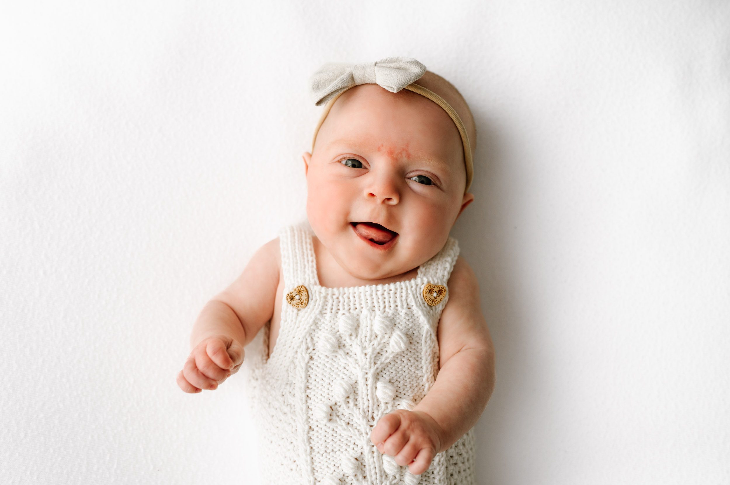 a baby girl wearing a white textured knit romper laying on a white backdrop and smiling up at the camera with her tongue sticking out