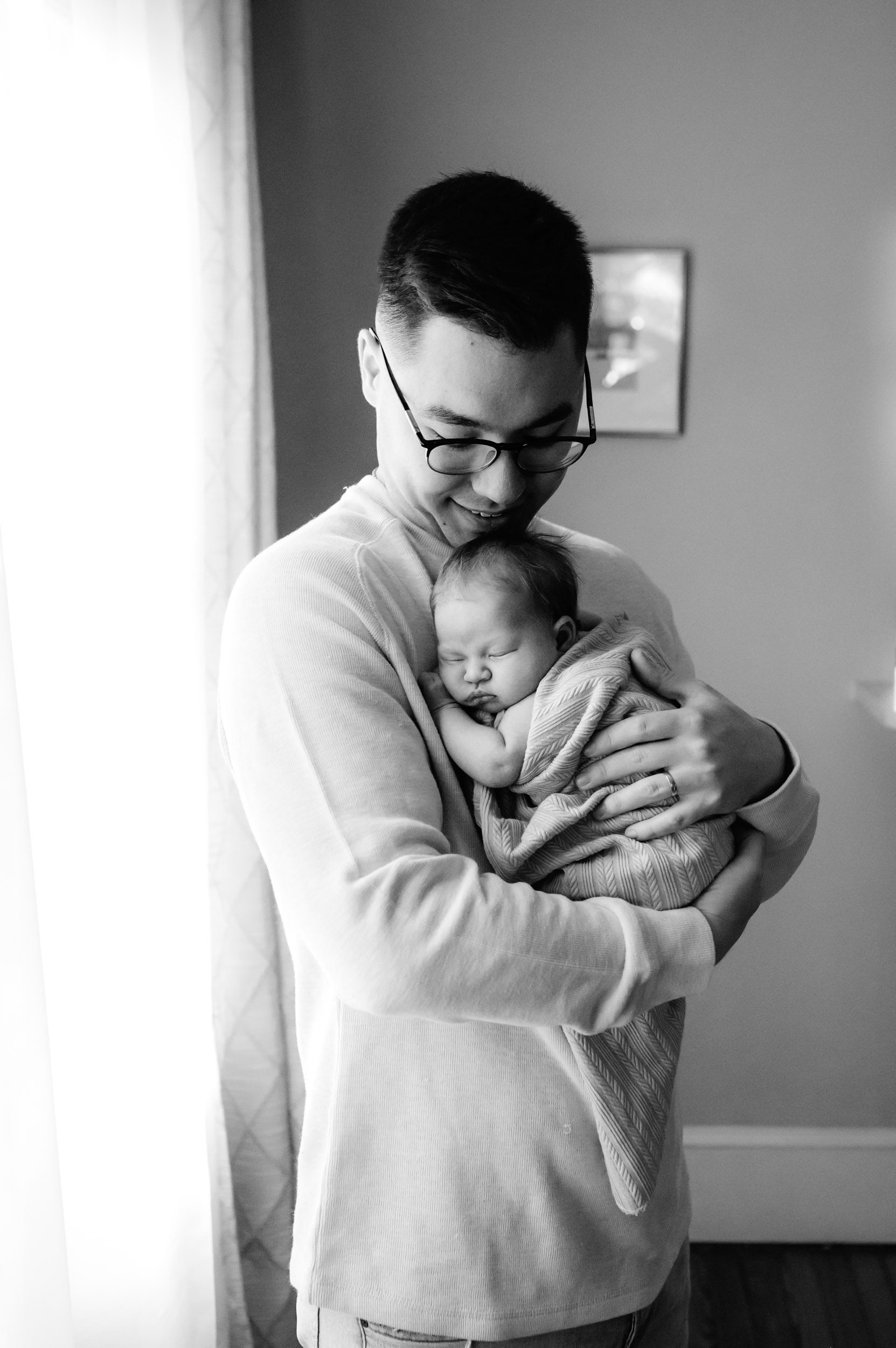 a black and white picture of a new father holding his baby girl against his chest and smiling down at her as she sleeps peacefully taken during an in home newborn photoshoot