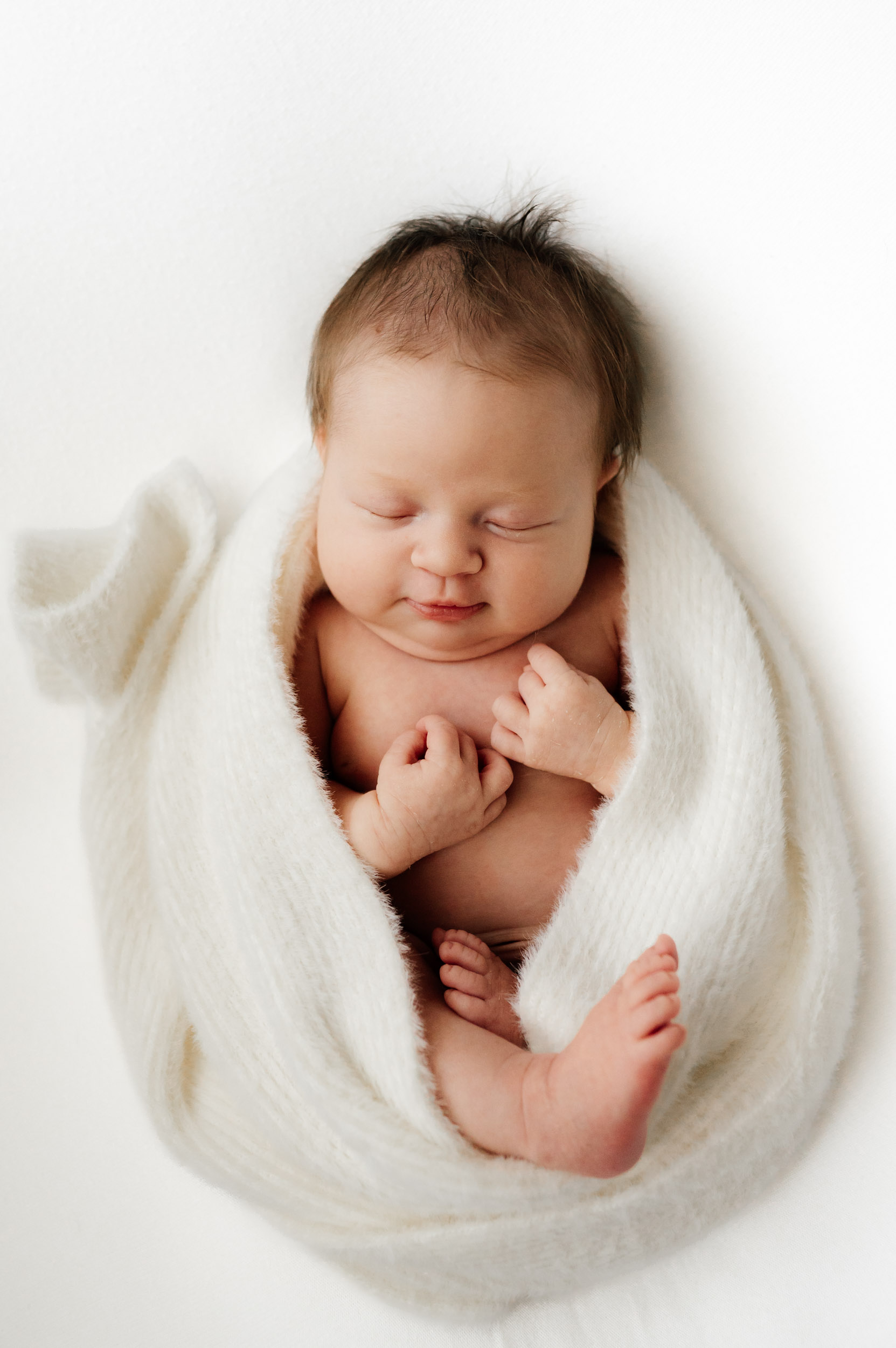 a baby girl laying on a white backdrop wrapped in a fuzzy white wrap with her hands and feet tucked in close to her body