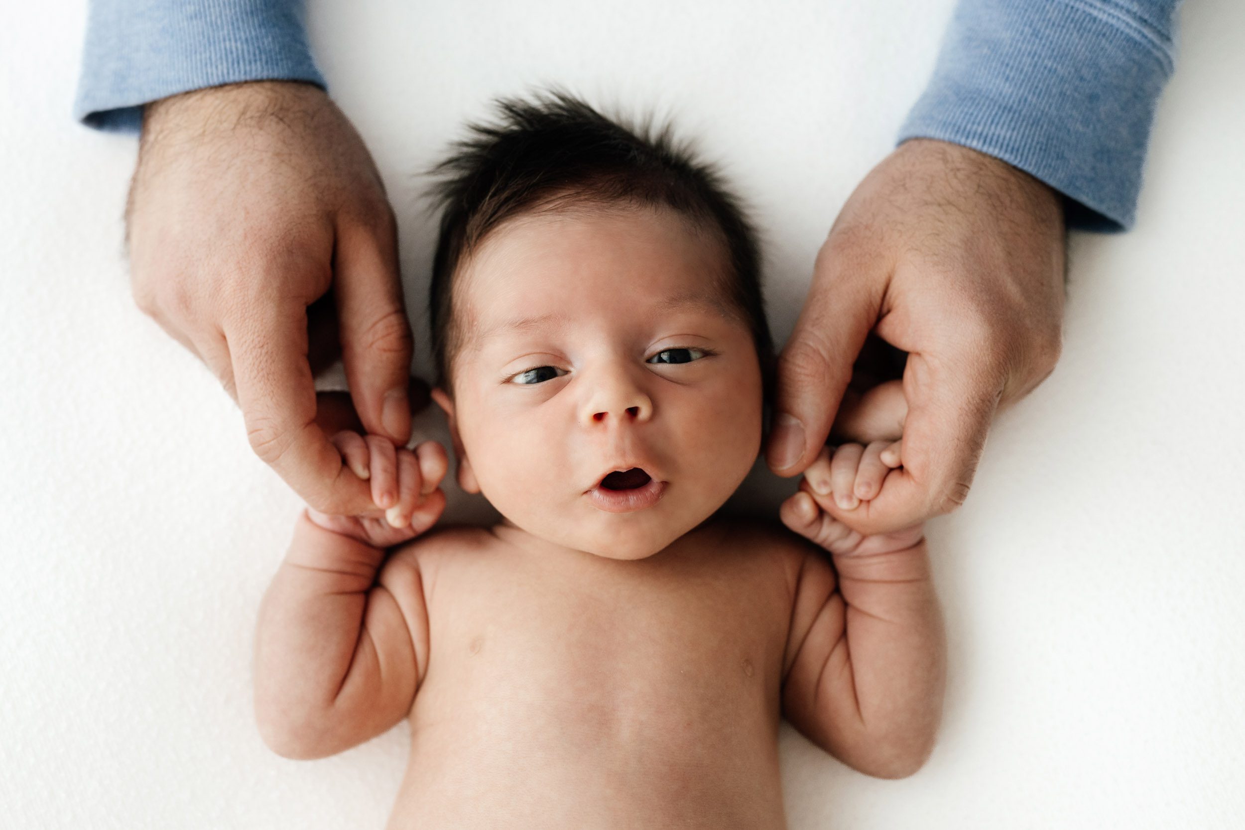 a baby boy laying on a white backdrop and grabbing onto his dad's fingers with his hands as he looks right at the camera during a natural newborn photoshoot