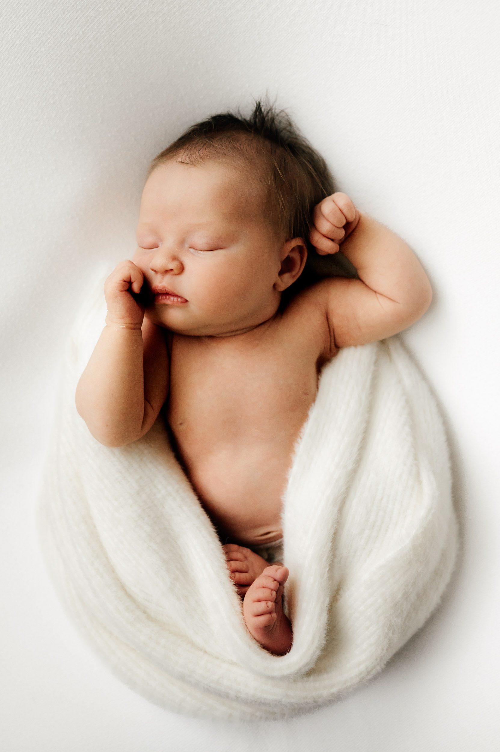 a baby girl wrapped in a white fuzzy blanket laying on a white backdrop sleeping with her hands resting against each side of her head during an in home newborn photoshoot