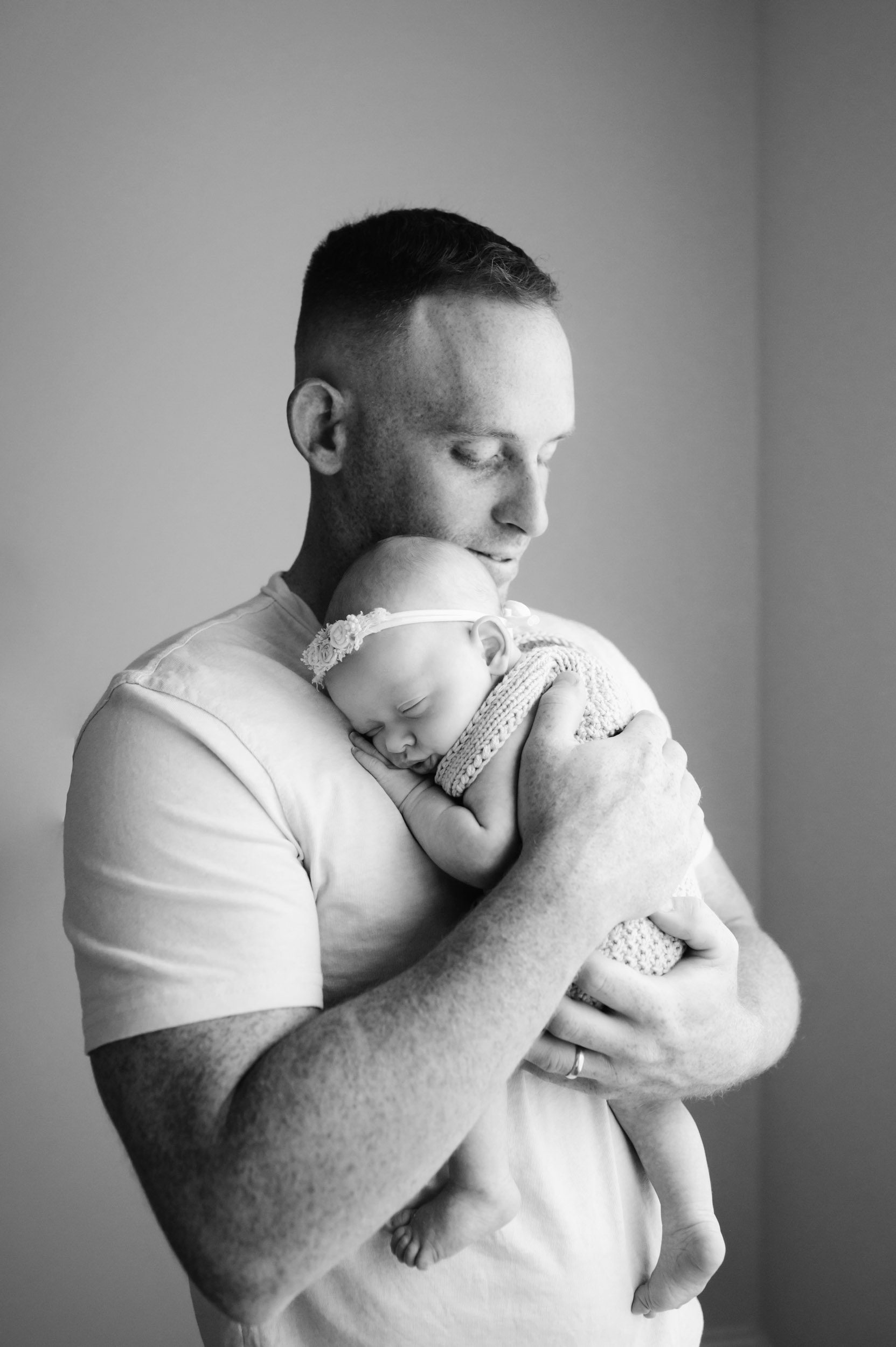 a black and white picture of a baby girl snuggled up against her dad's chest sleeping as he smiles down at her during an in home newborn photography session