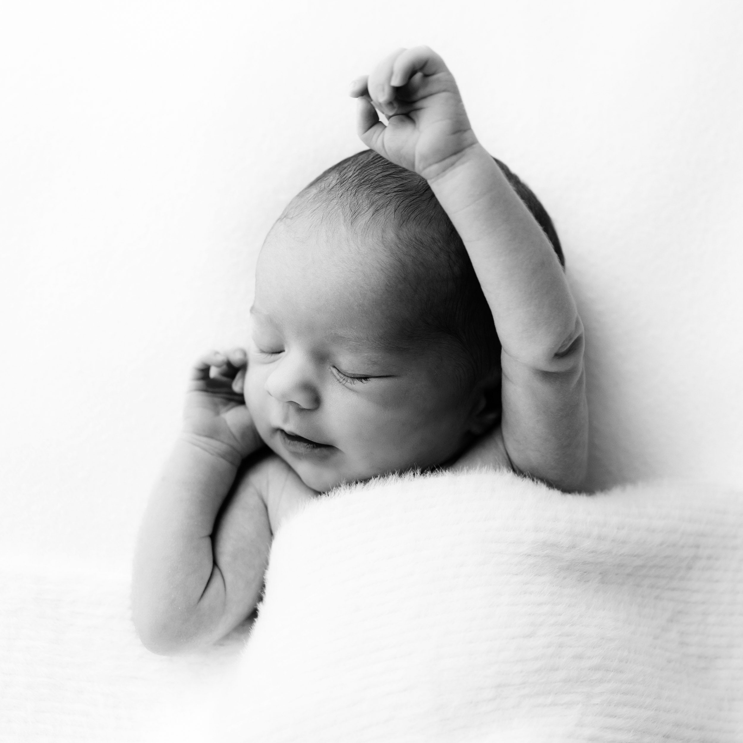 a black and white picture of a baby girl laying on a white backdrop under a fuzzy white blanket and stretching her arms as she sleeps during a newborn photo session