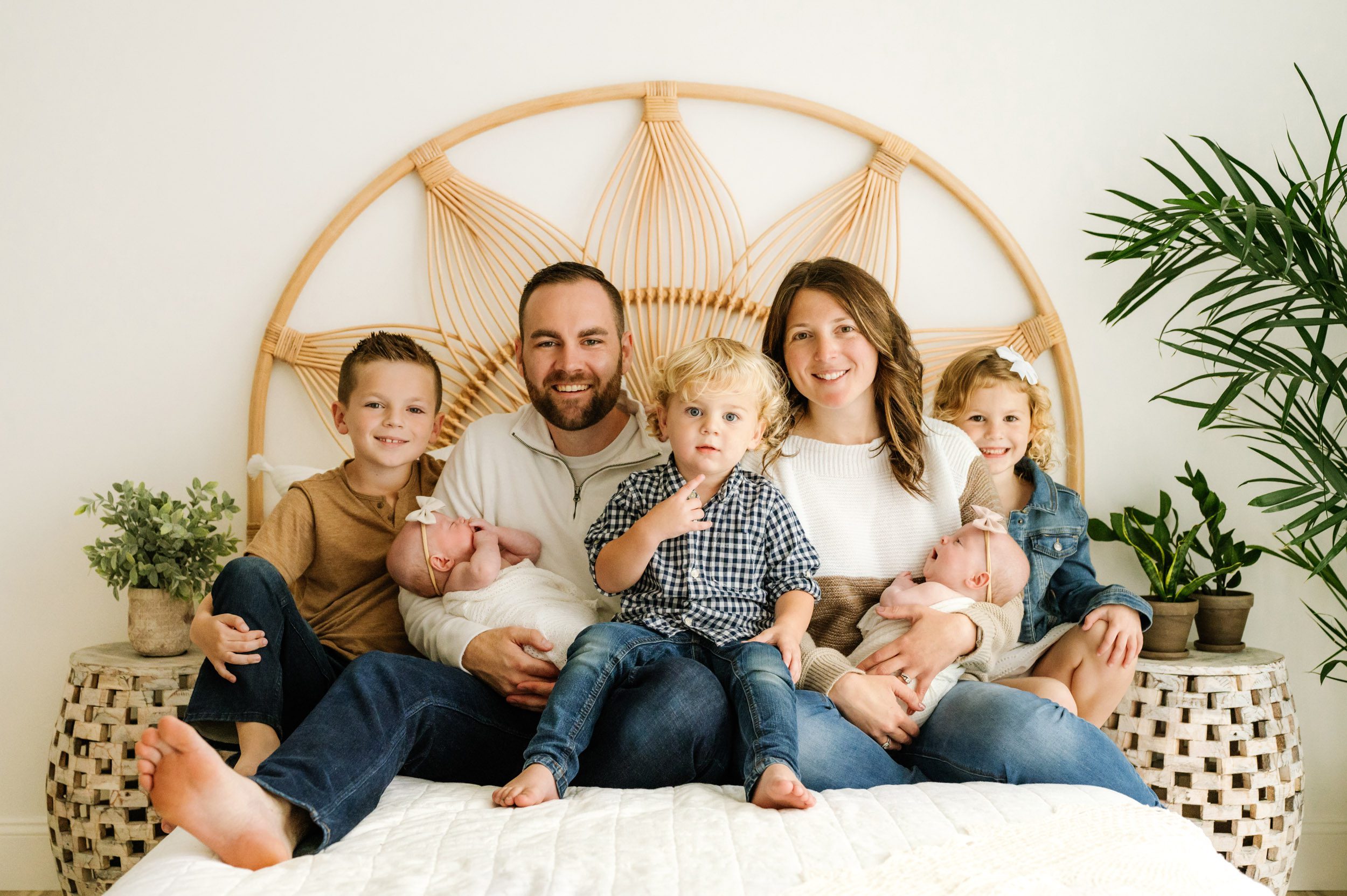 a family of seven sitting on a bed and smiling at the camera while mom and dad each cradle one of their twin baby girls in their arms during a lifestyle newborn photo session