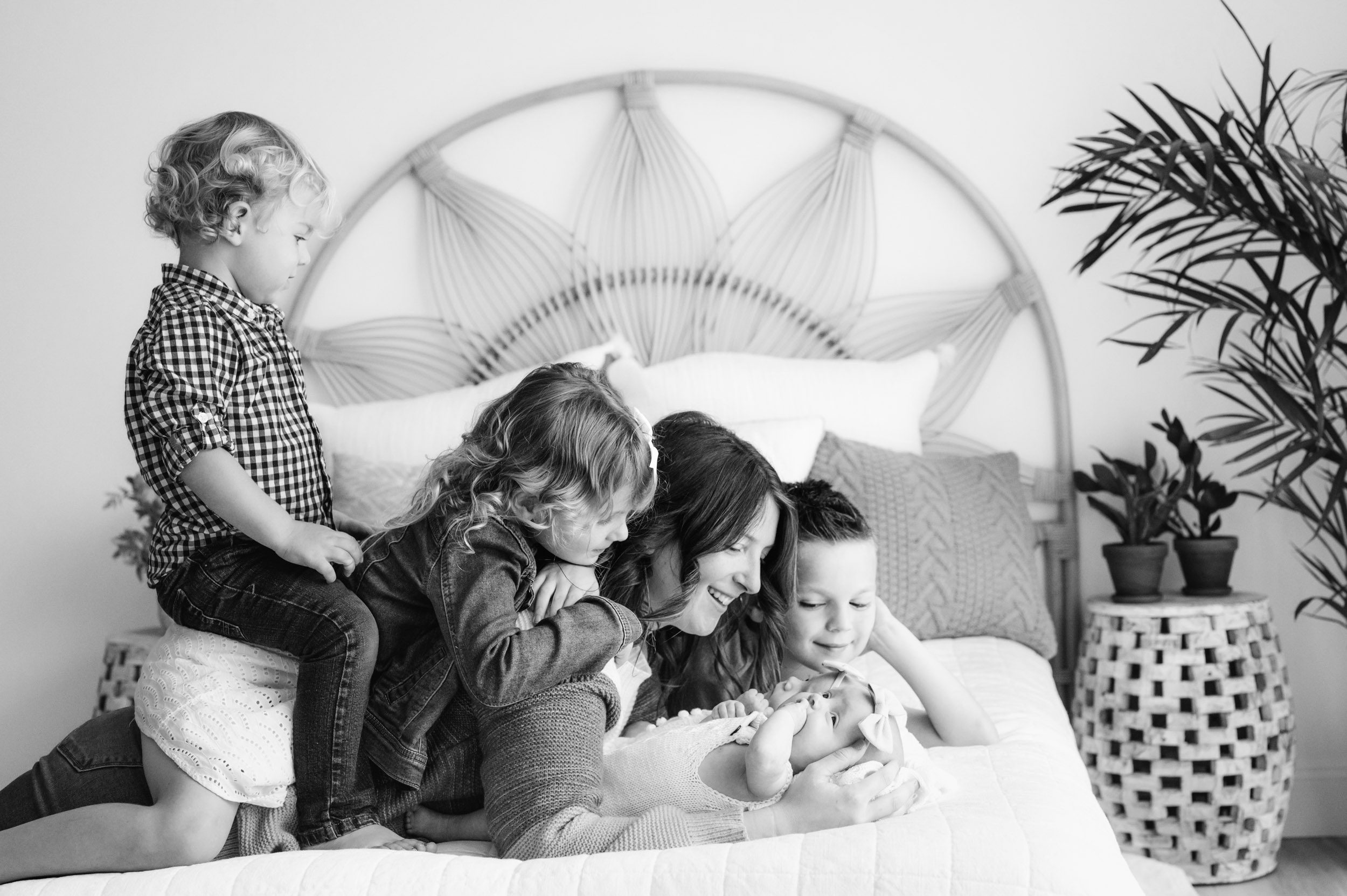 a black and white picture of a mom laying on a bed with her twin baby girls and her three older kids all snuggled around her as they all smile down at the babies during a lifestyle newborn photoshoot