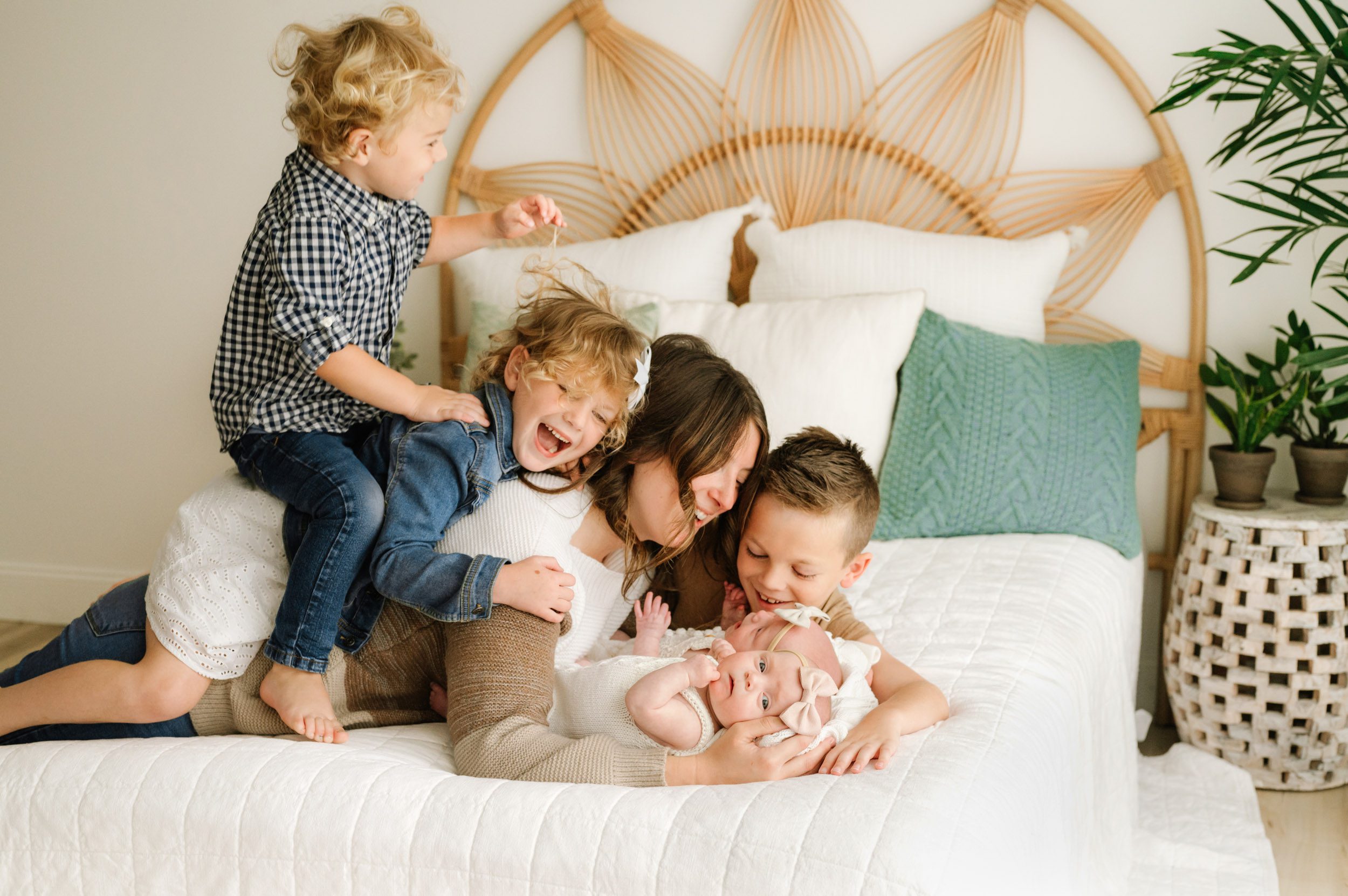 a mom laying on a bed with her twin baby girls as her oldest son lays next to her, her daughter lays on her back, and her youngest son sits on his sister's back and jumps up and down while they all laugh during a lifestyle newborn photoshoot