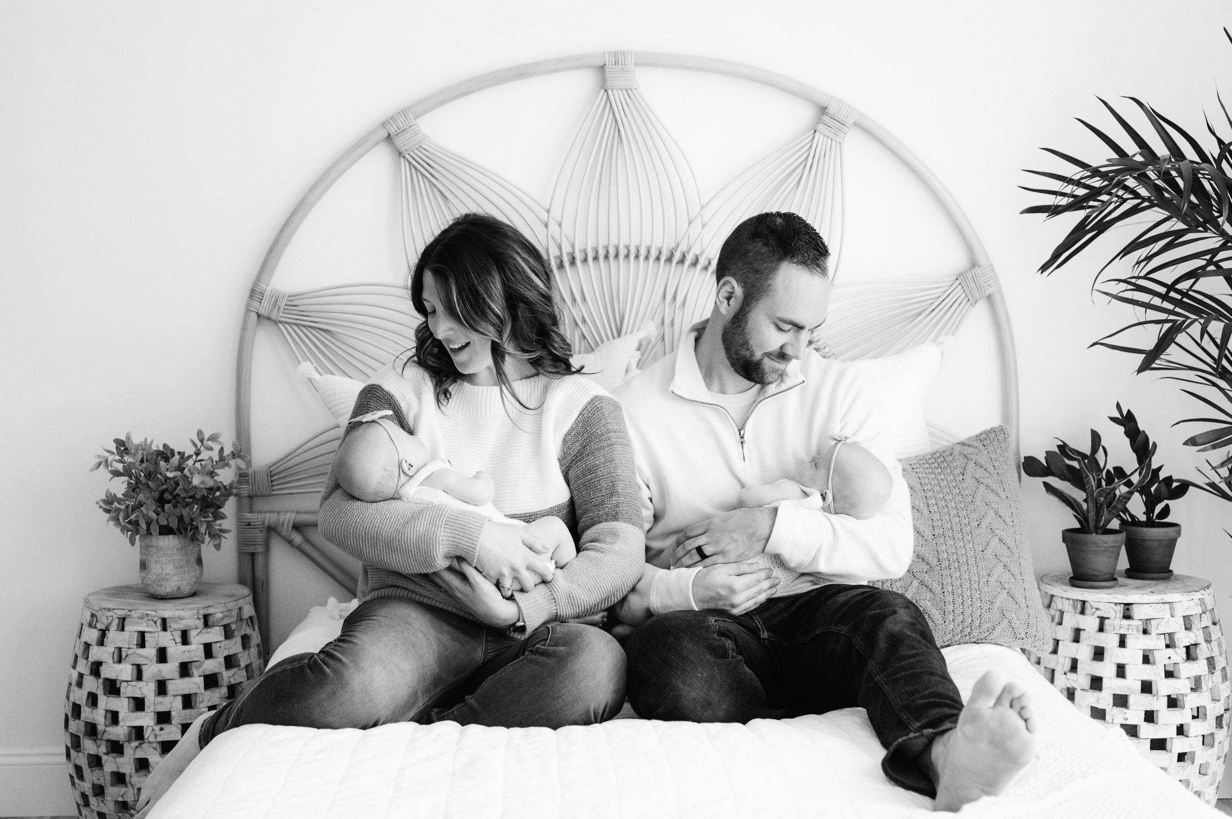 a black and white picture of new parents sitting on a bed while mom and dad each cradle one of their twin baby girls in their arms and smile down at them during a lifestyle newborn photo session